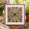 Framed cross-stitched artwork with floral patterns on a wooden table with plants and a teacup.