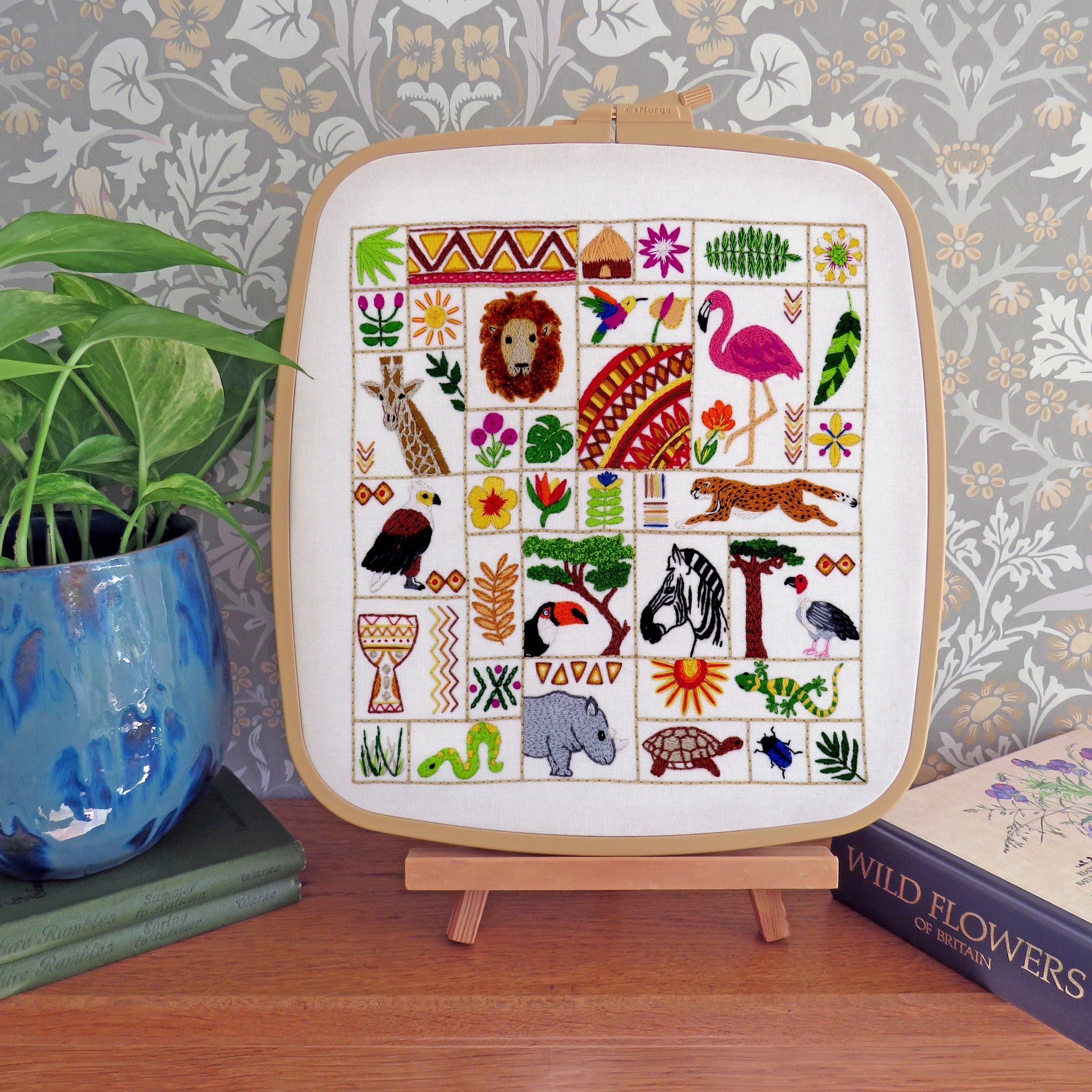 A square framed cross-stitch embroidery featuring colorful safari animal and nature motifs, displayed on a small wooden stand on a wooden table with a potted plant and a book in the background.