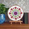 Embroidery hoop with floral pattern on a wooden stand against a decorative wall.