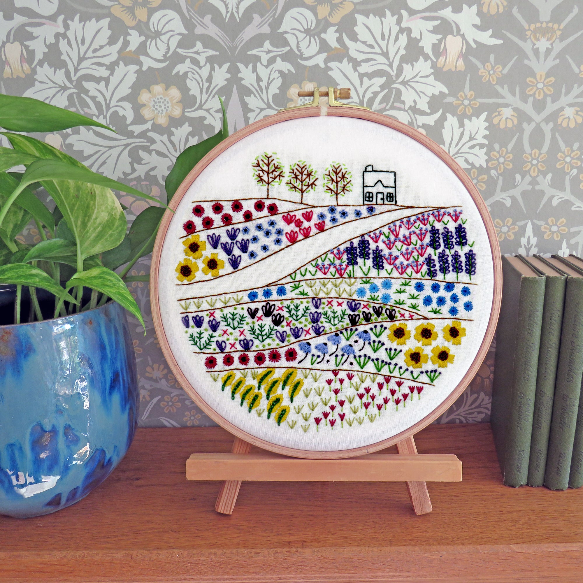 A circular embroidery hoop displaying a colorful embroidered landscape with hills, flowers, trees, and a small house, shown on a small wooden stand on a tabletop with a potted plant and books in the background.