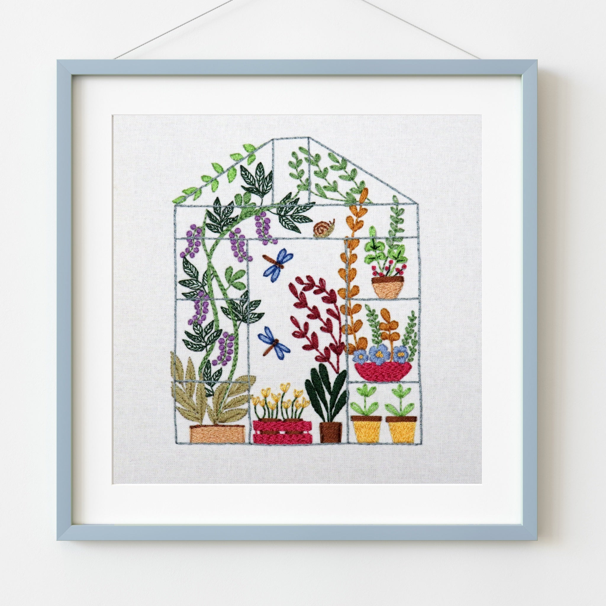 Embroidered artwork of a greenhouse with plants and flowers in pots, framed in light blue.