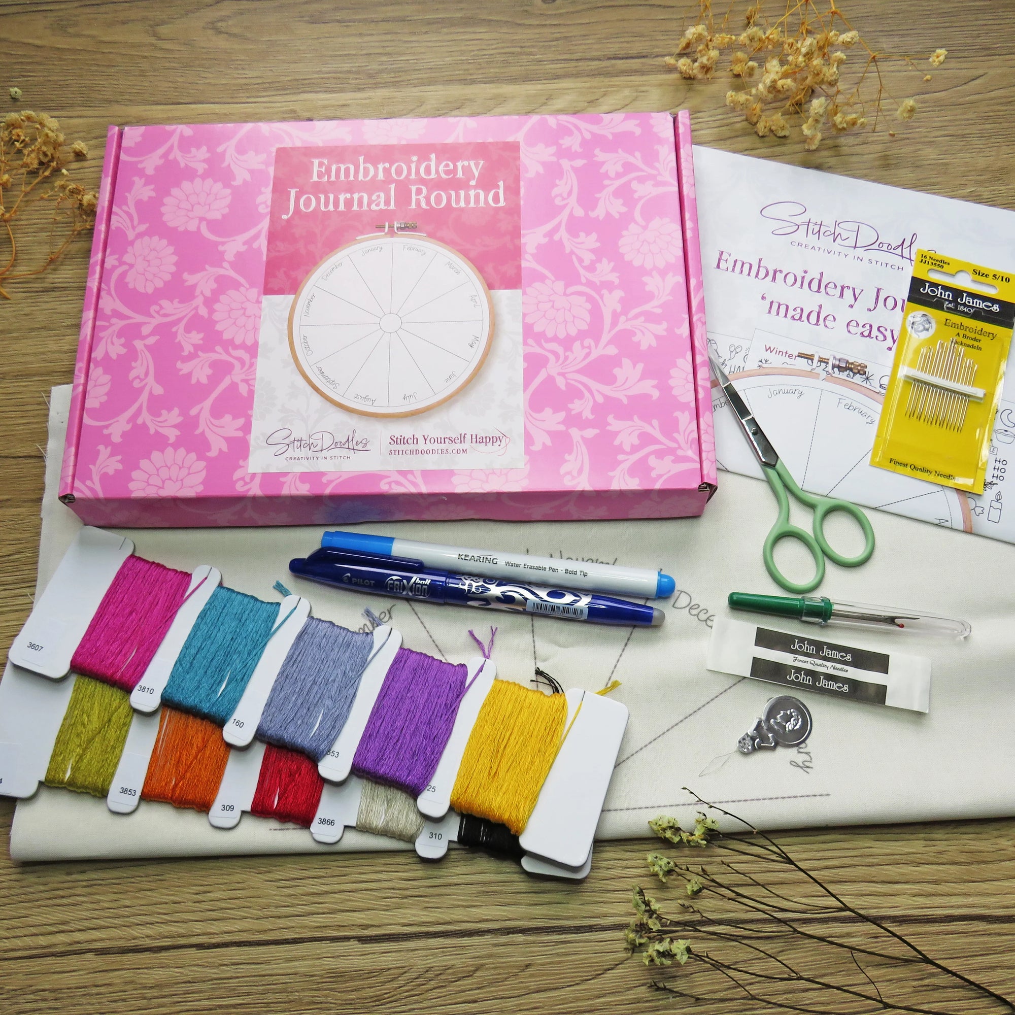 Embroidery kit with pink box, threads, pens, and scissors on a wooden surface.