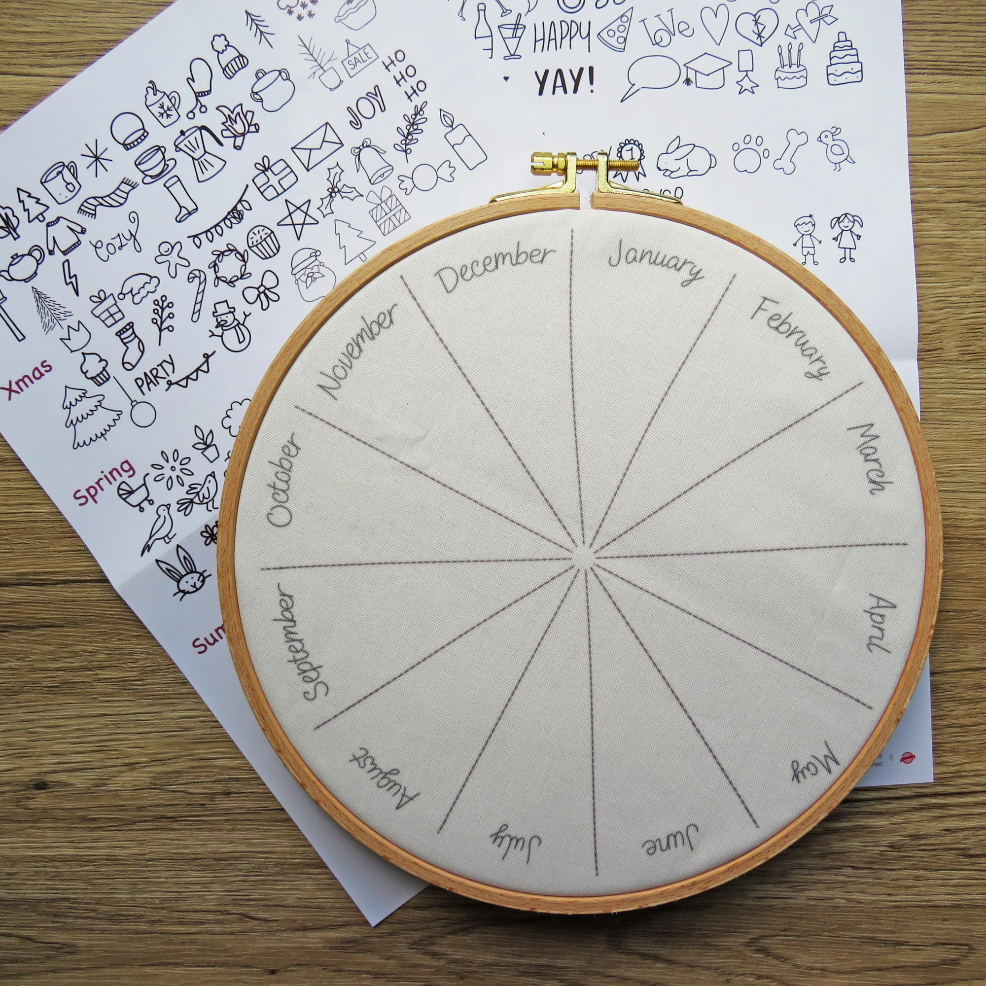 Embroidery hoop with a calendar design on fabric, placed on a wooden surface with a sheet of doodles.