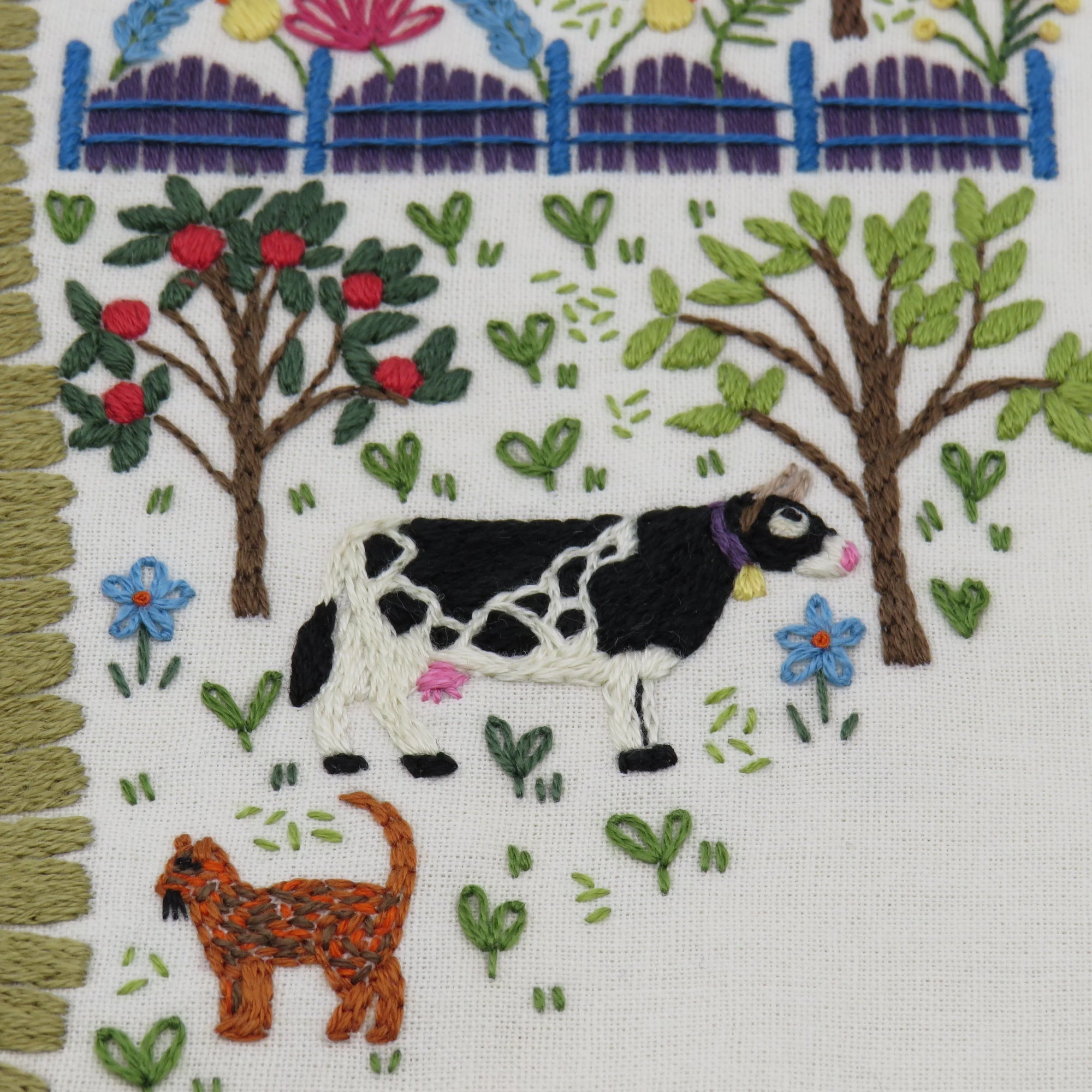 Embroidered scene with a cow and cat in a garden setting.