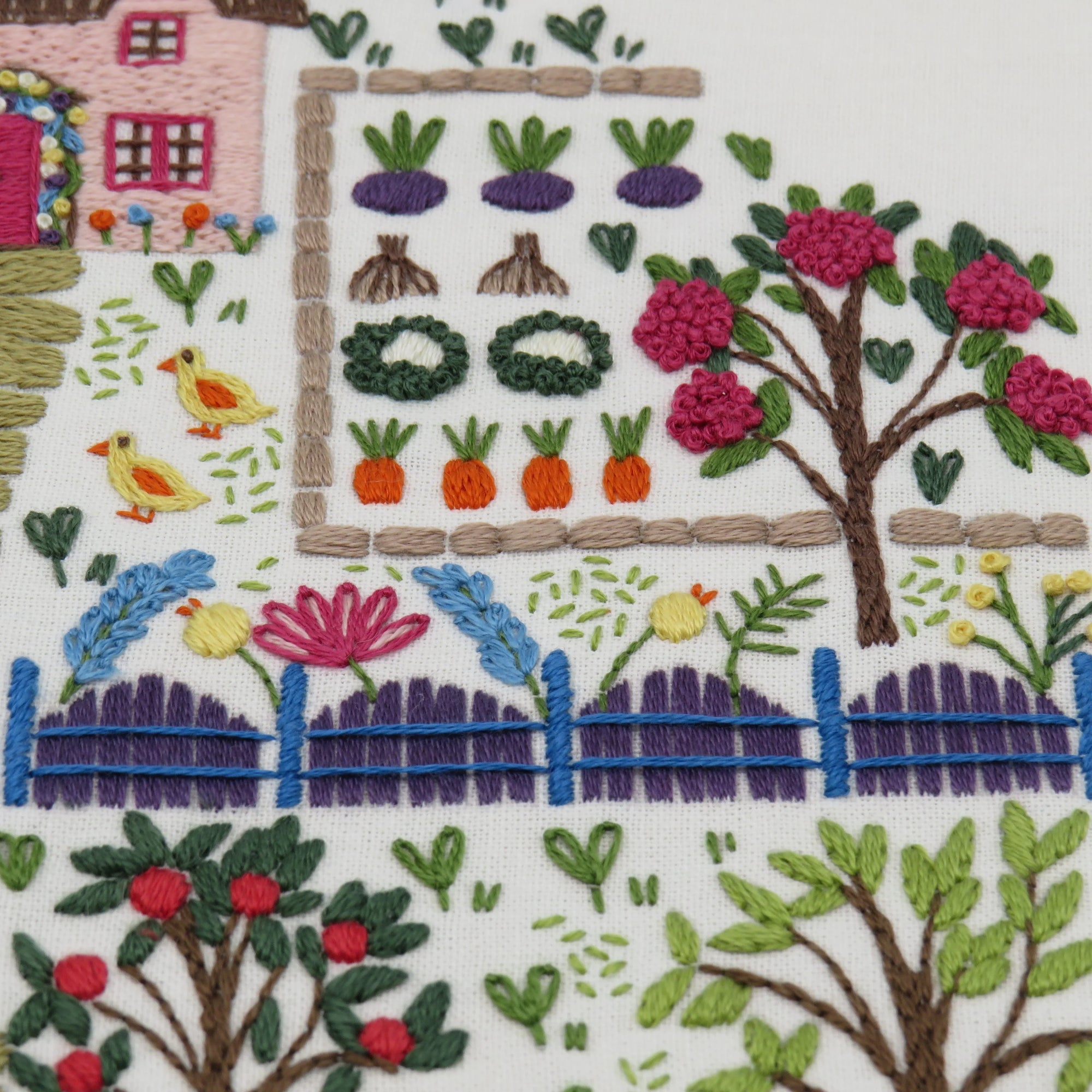 Embroidered scene of a garden with flowers, vegetables, and a house.