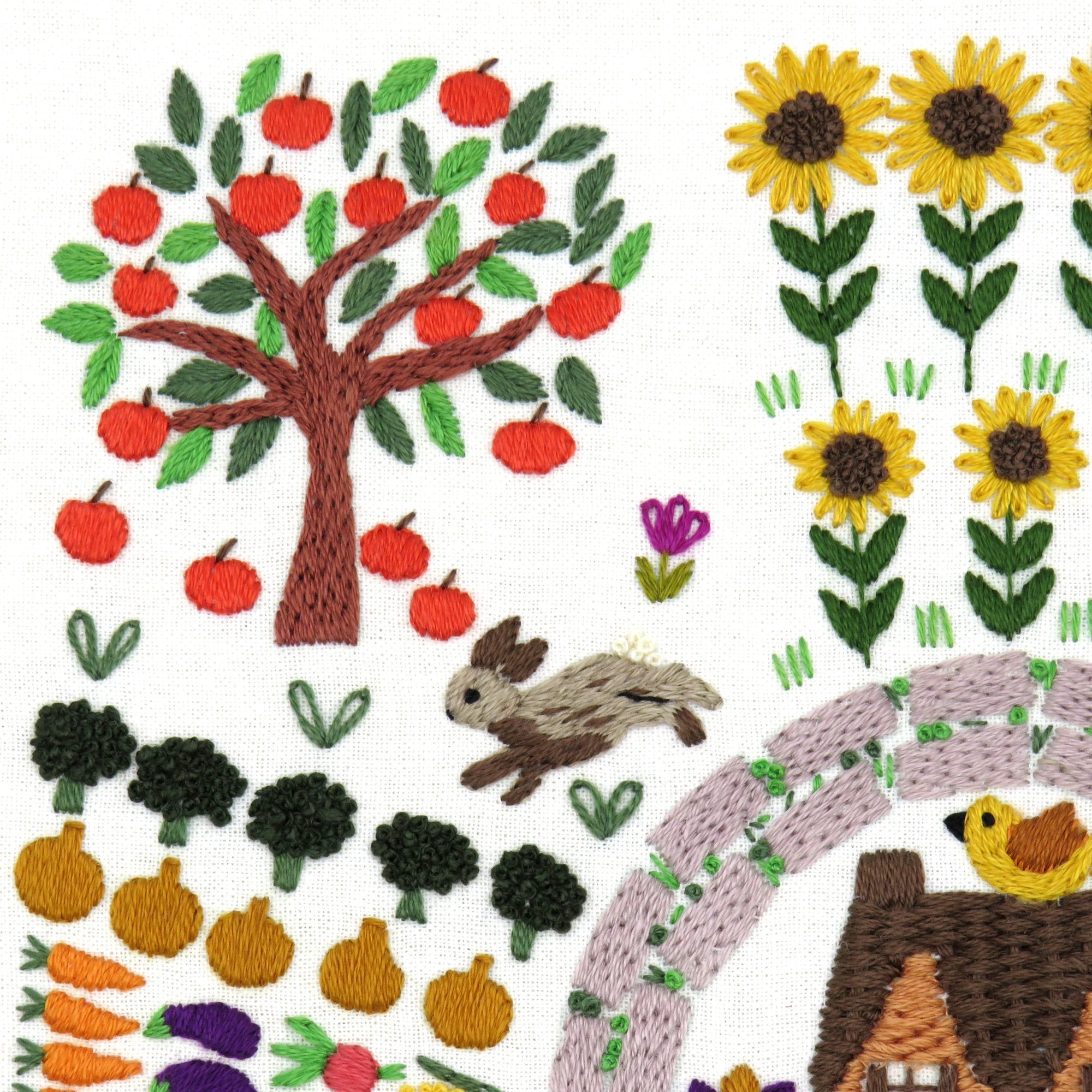 Embroidered design of a garden scene with trees, flowers, animals, and vegetables.