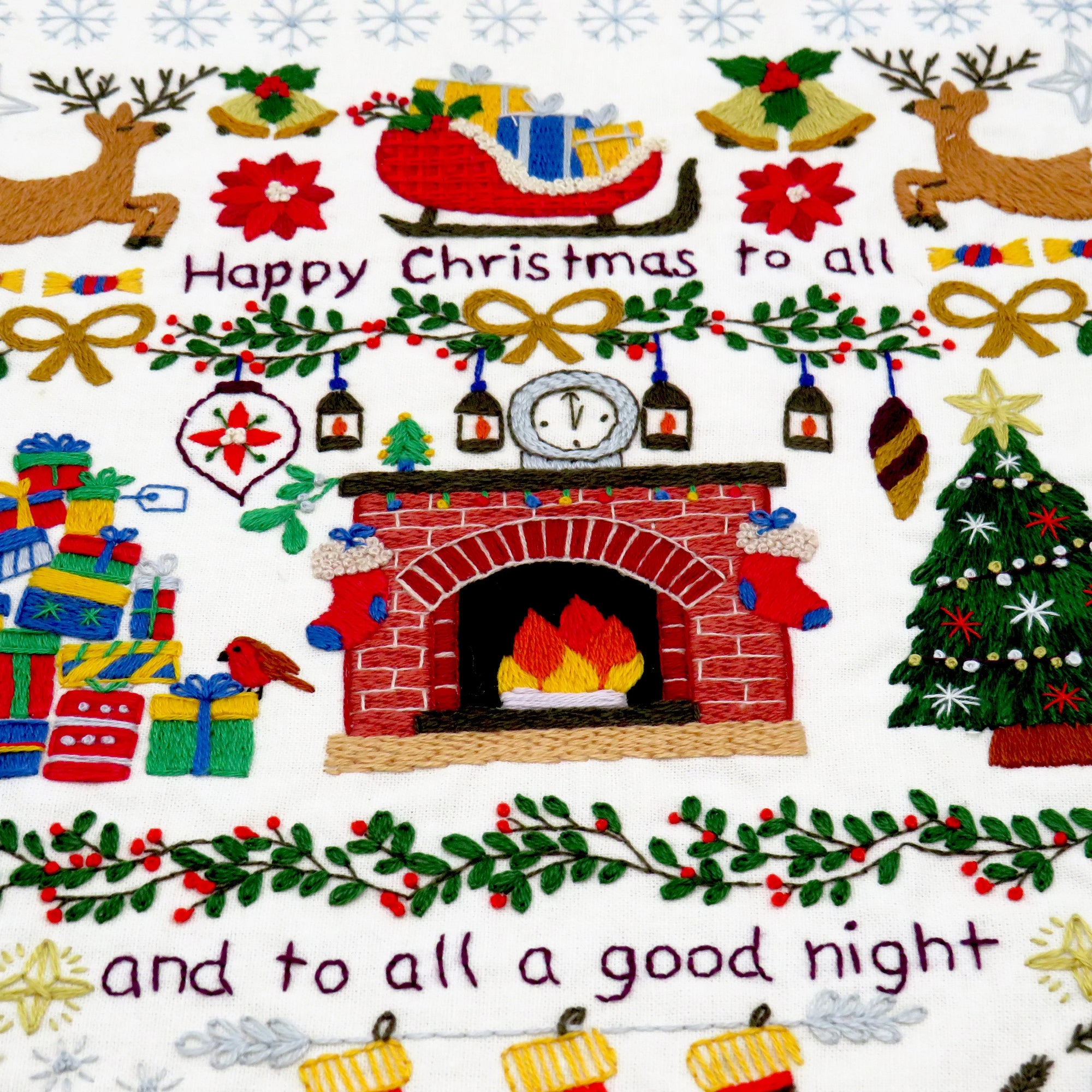 Embroidered Christmas scene with fireplace, reindeer, presents, and text on a white background.