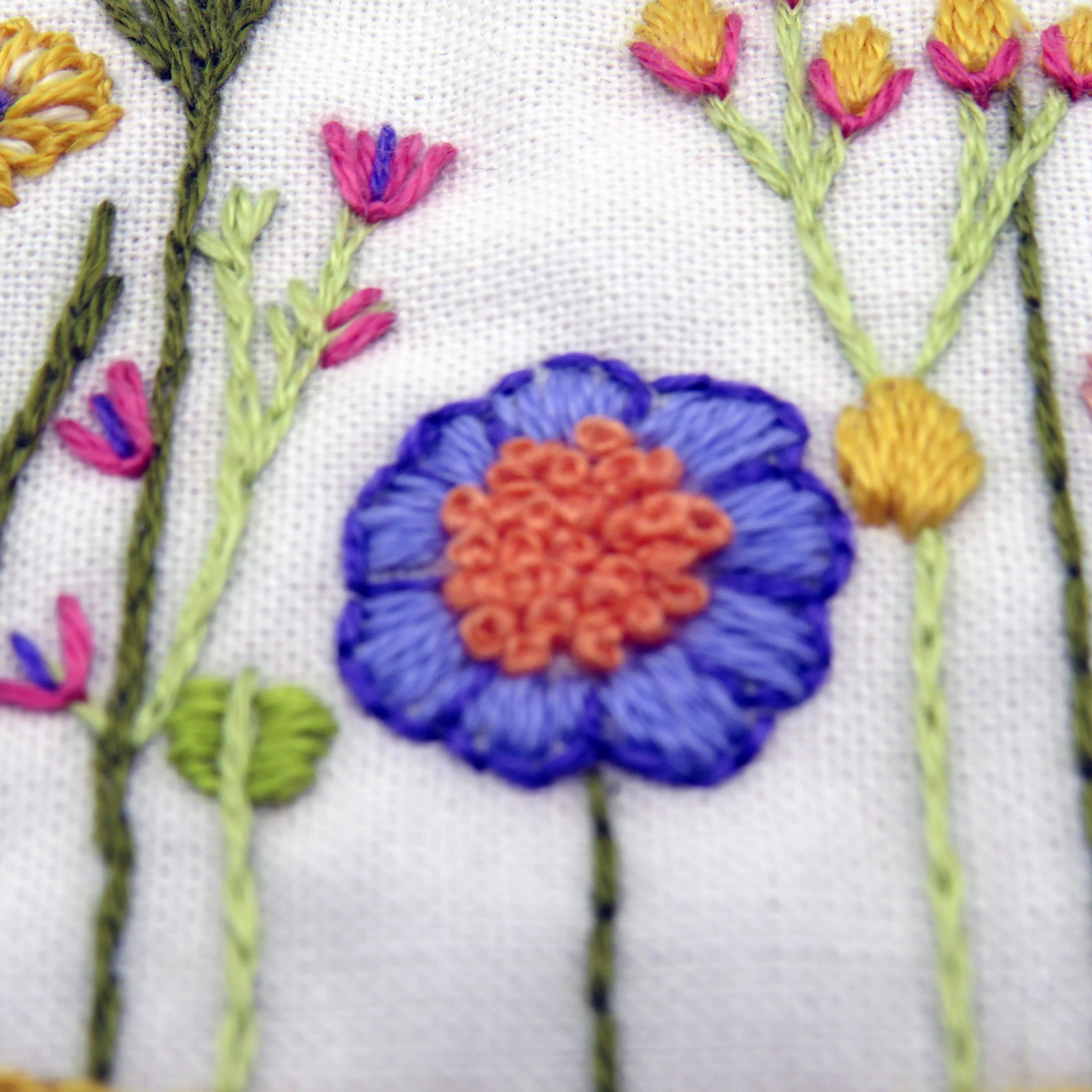 Embroidered floral design with a blue flower and orange center on white fabric.
