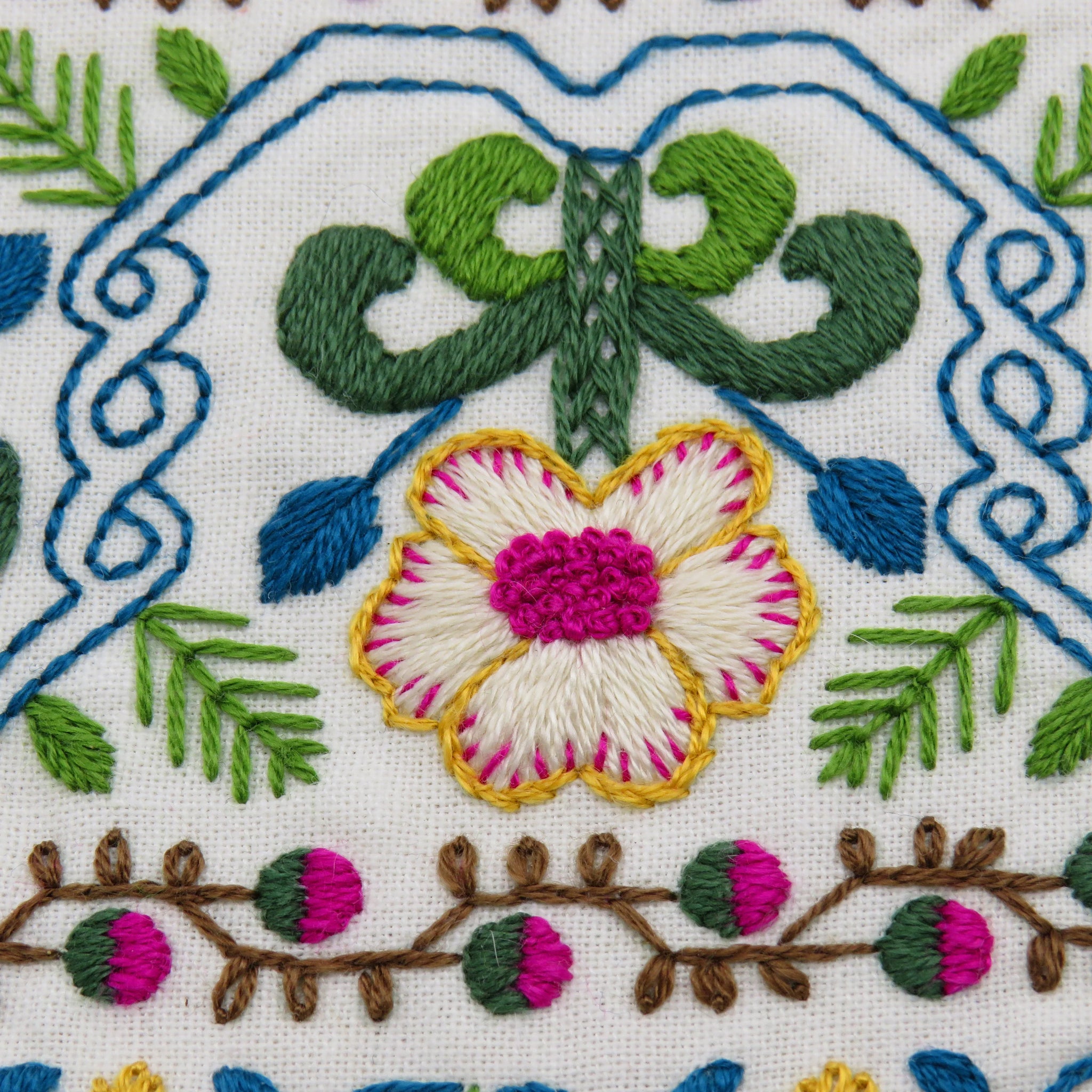 Hand embroidery pattern featuring a vibrant flower with pink center, green leaves, and blue vines on cream fabric.