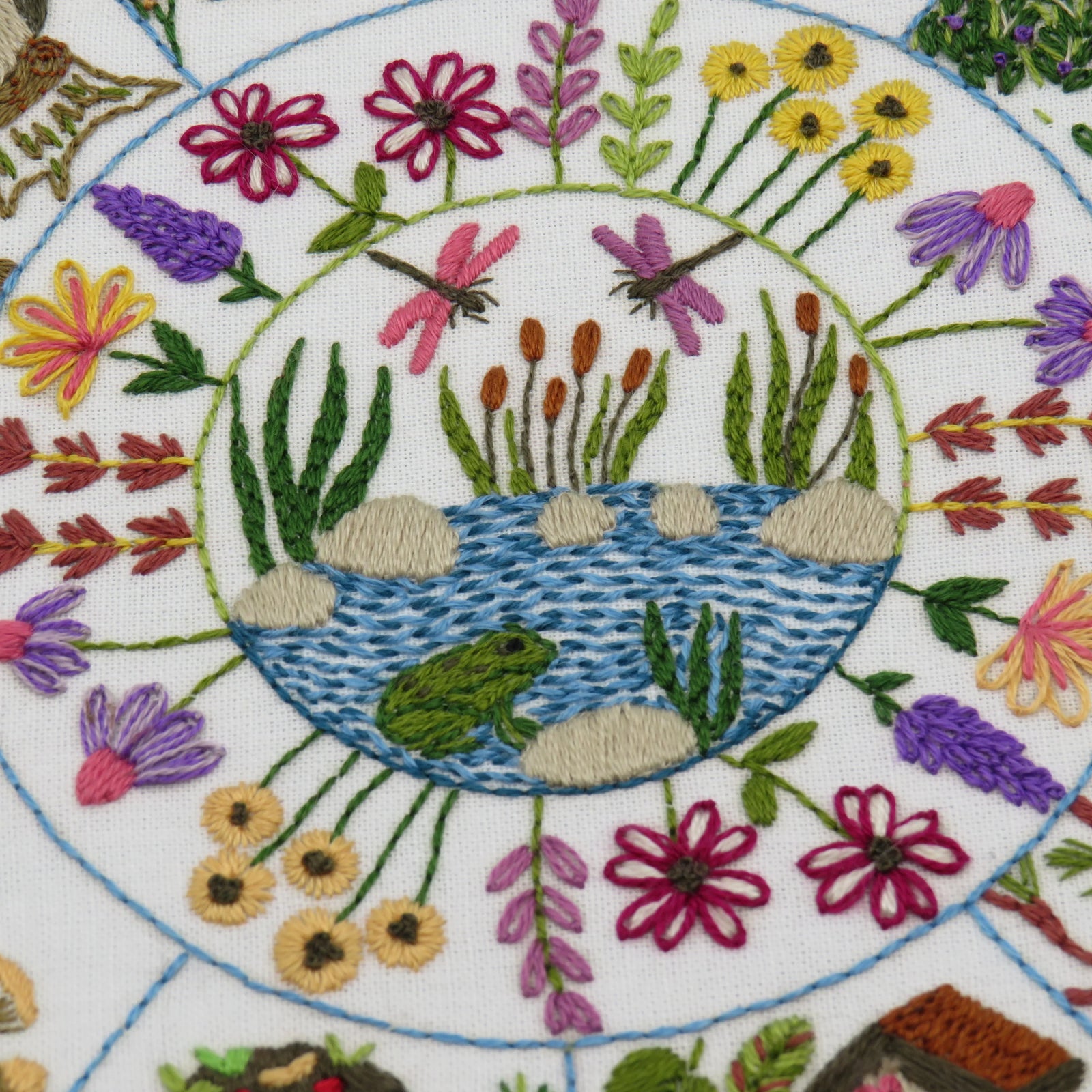 Embroidered design of a pond with a frog, surrounded by flowers and plants on a white background.