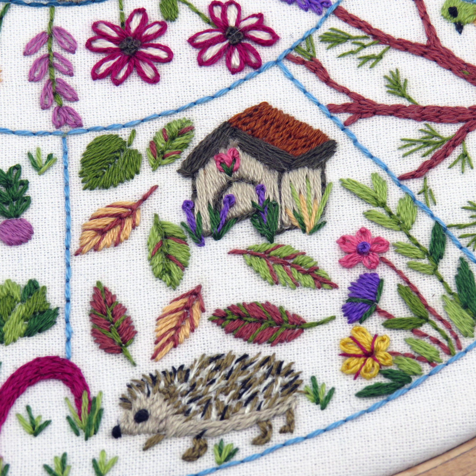 Embroidered design with a hedgehog, flower house, and foliage on white fabric.