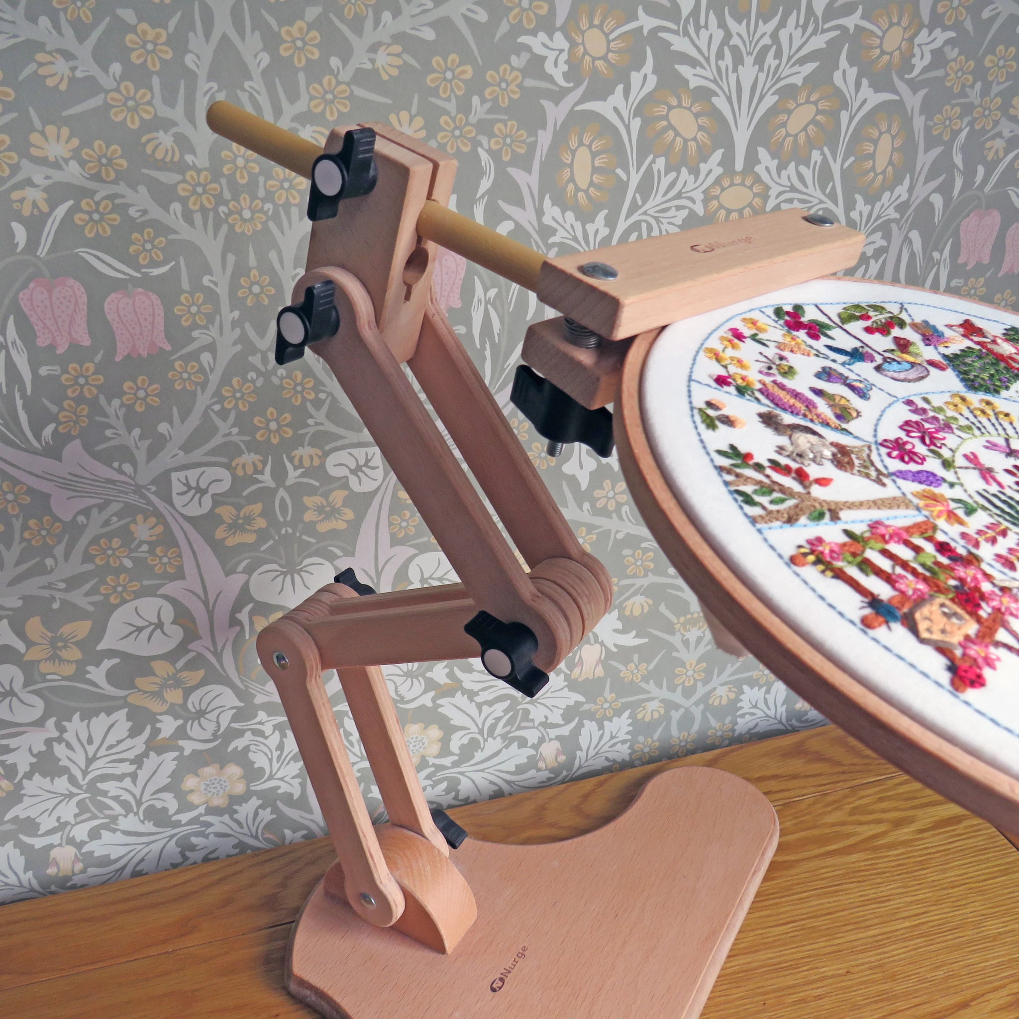 Wooden embroidery hoop stand with an embroidery hoop containing a colorful design on a wooden table.