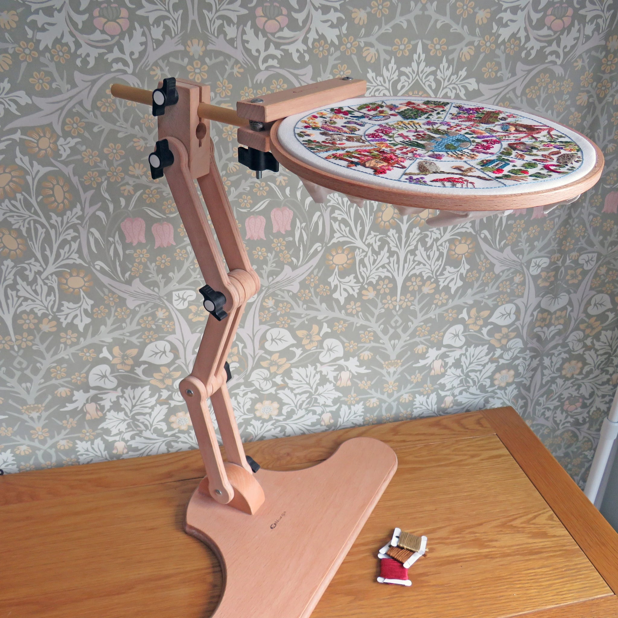 Wooden embroidery hoop stand holding an embroidered piece on a wooden table with floral wallpaper.