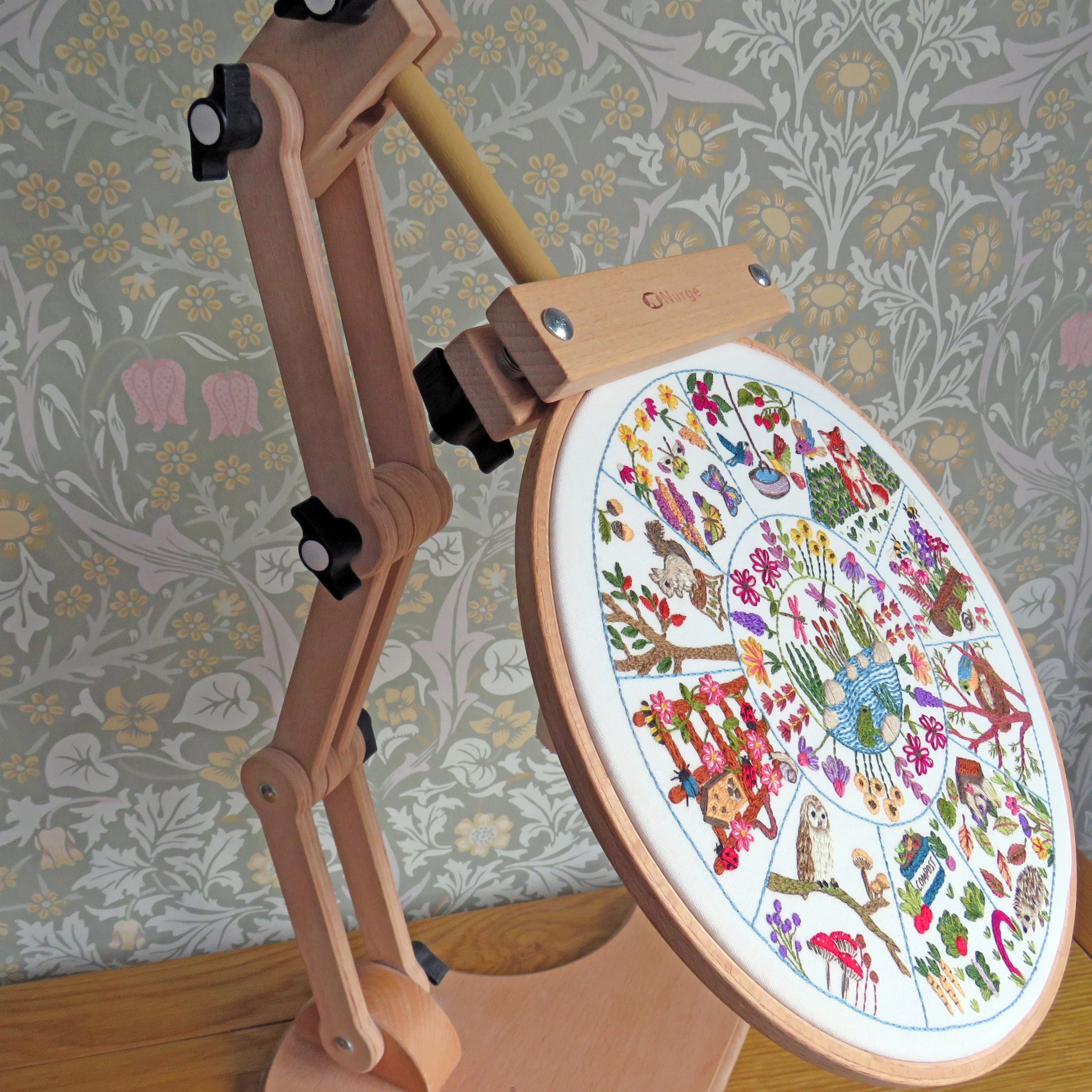 Embroidery hoop with colorful design on a stand against a floral wallpaper background