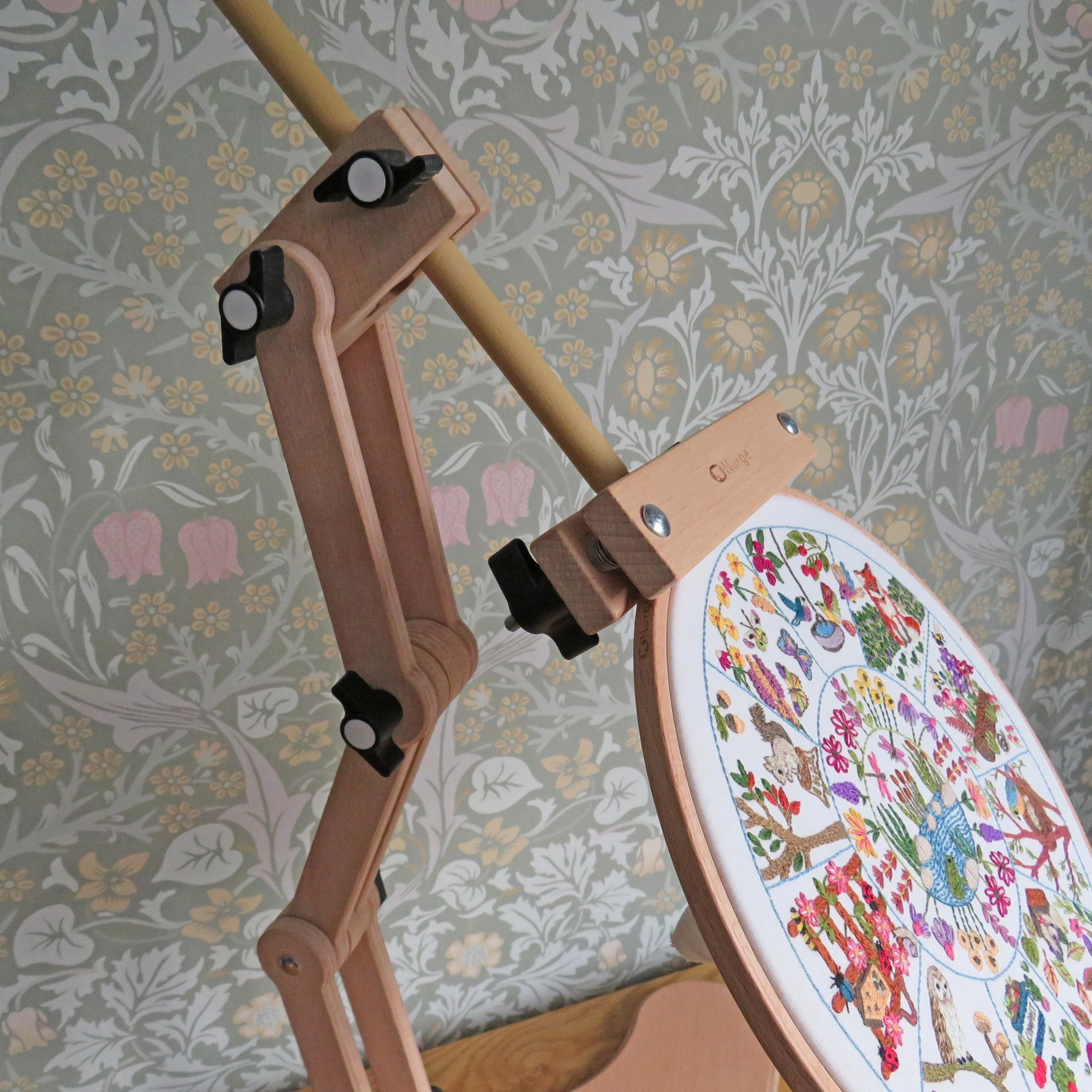 Wooden embroidery hoop with a colorful cross-stitched design against a floral wallpaper background.