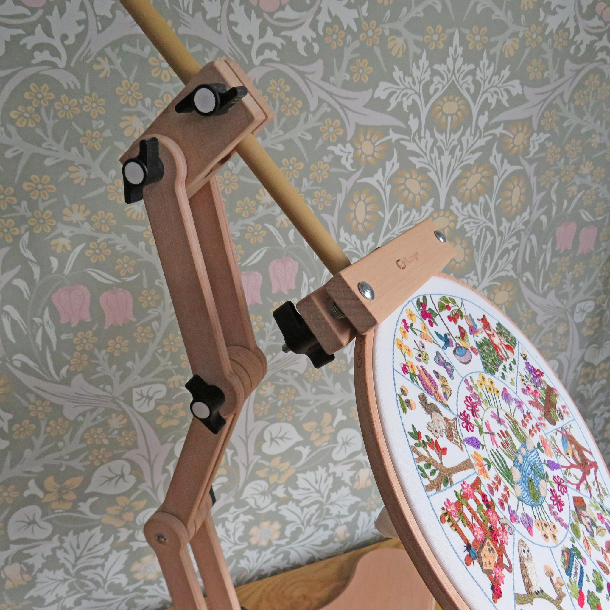 Wooden embroidery hoop with a colorful cross-stitched design against a floral wallpaper background.