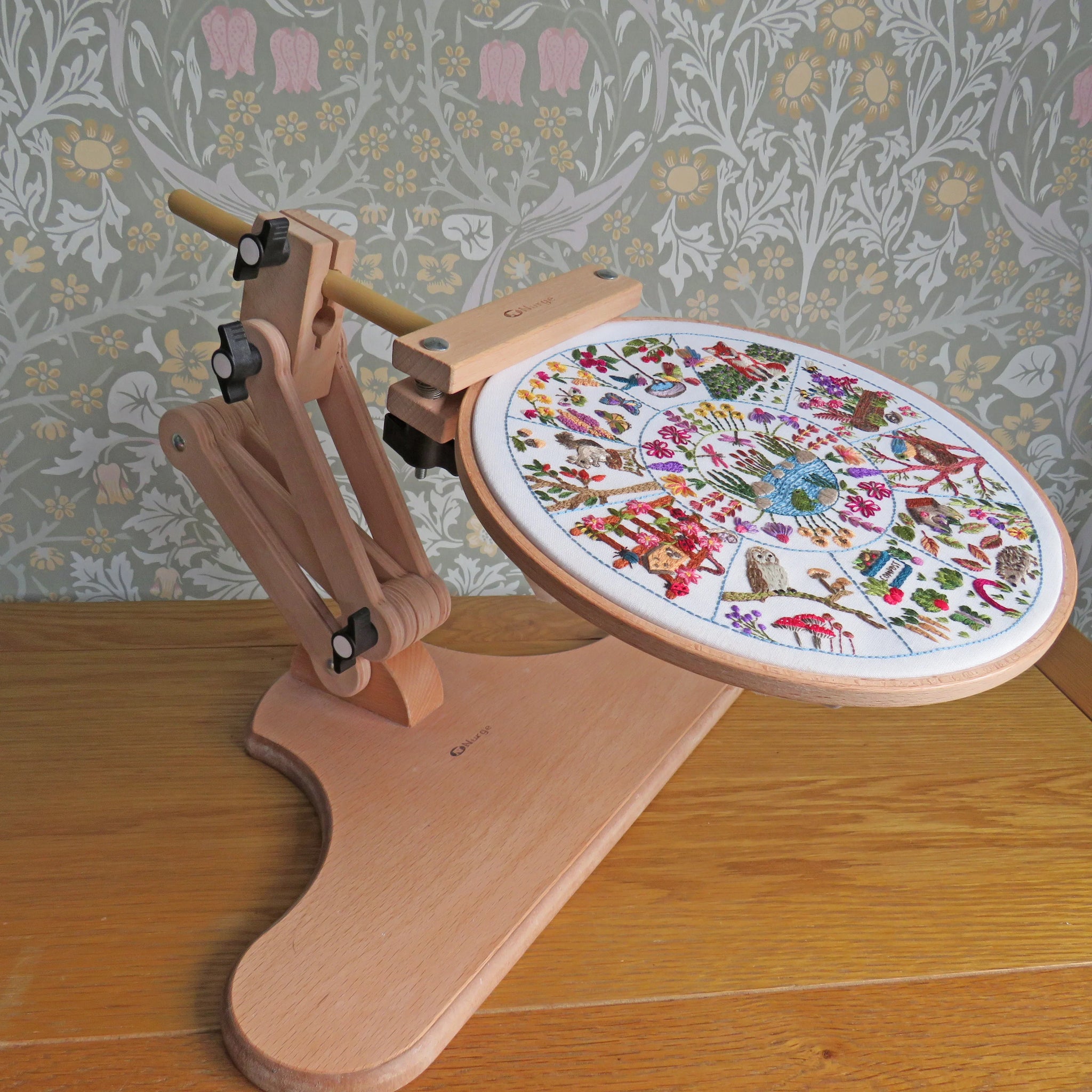 Embroidery hoop with a colorful embroidered design on a wooden stand against a floral wallpaper background.