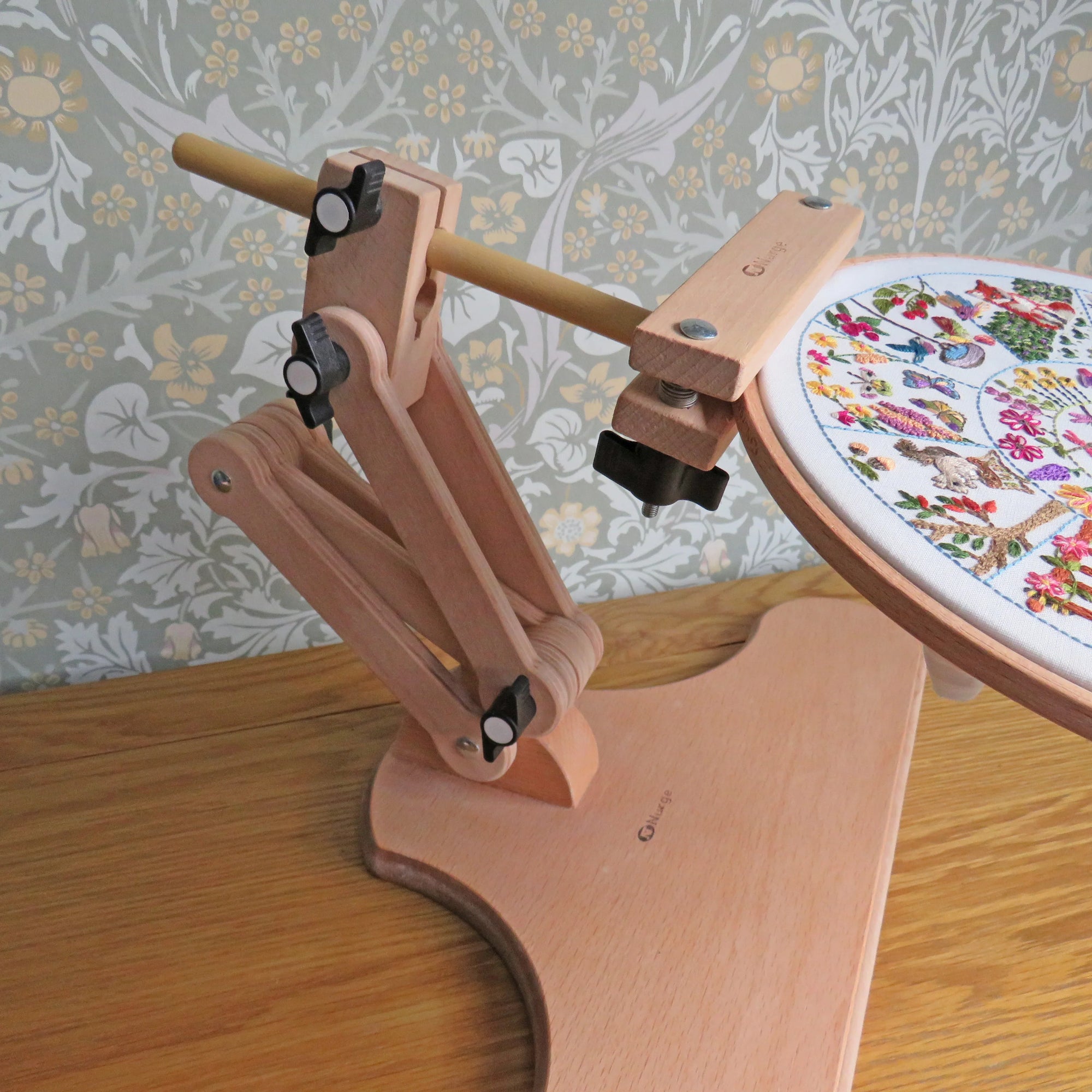 Wooden embroidery hoop stand with an embroidered piece on a wooden surface and floral wallpaper background.