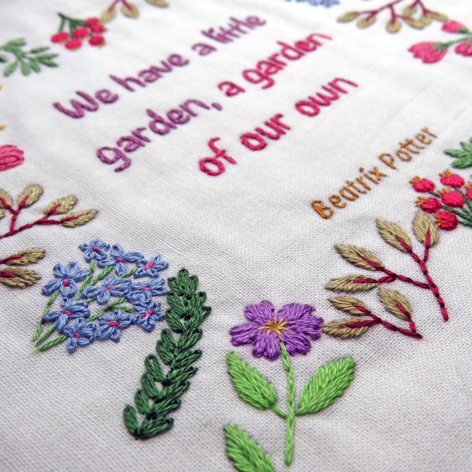 Embroidered text and floral designs on a white fabric with 'We have a little garden, a garden of our own' and 'Beatrix Potter' text.
