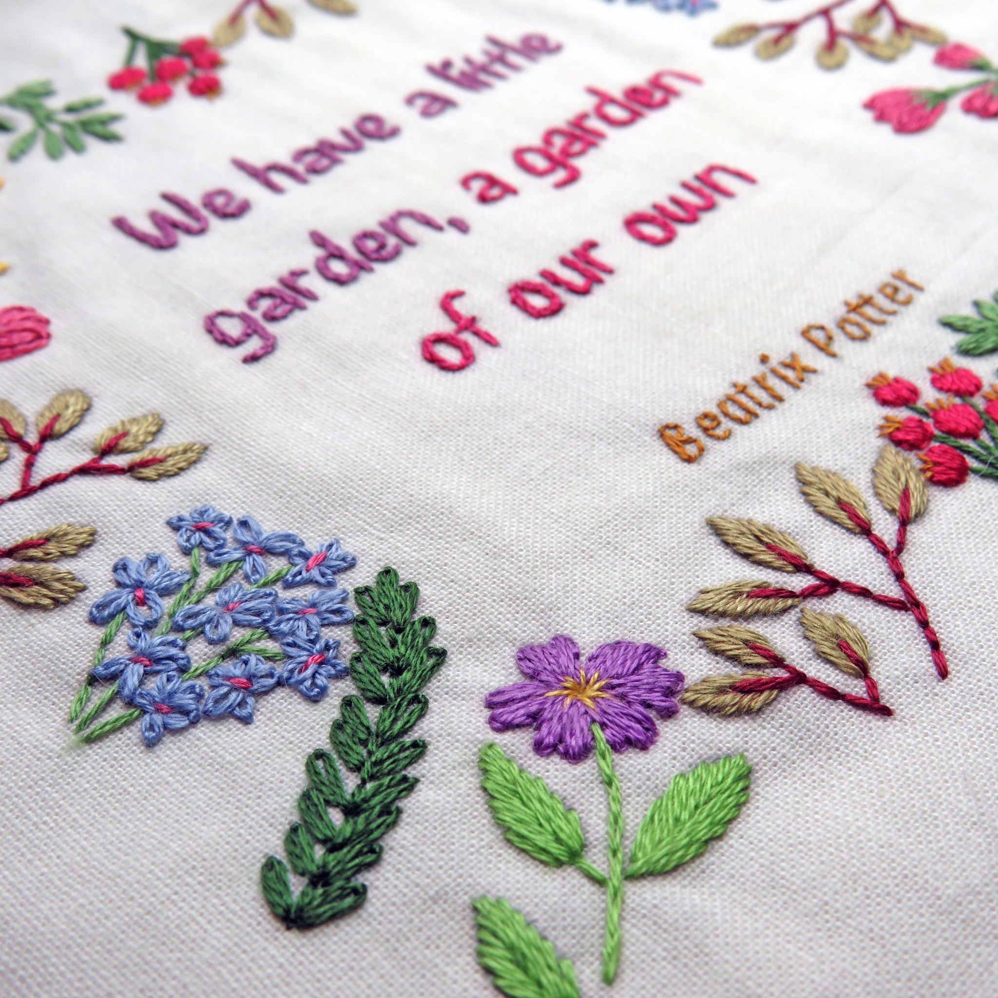 Embroidered text and floral designs on a white fabric with 'We have a little garden, a garden of our own' and 'Beatrix Potter' text.