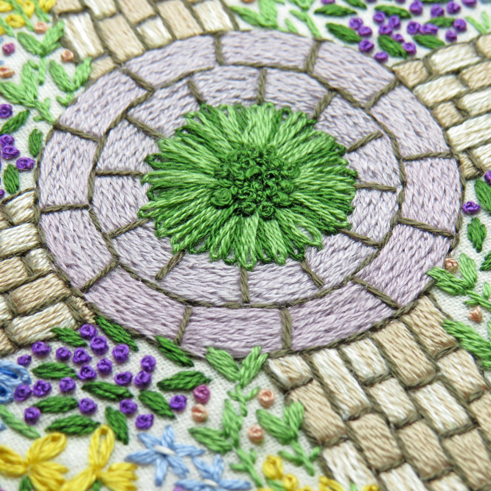 Embroidered design of a circular stone path with greenery and flowers.