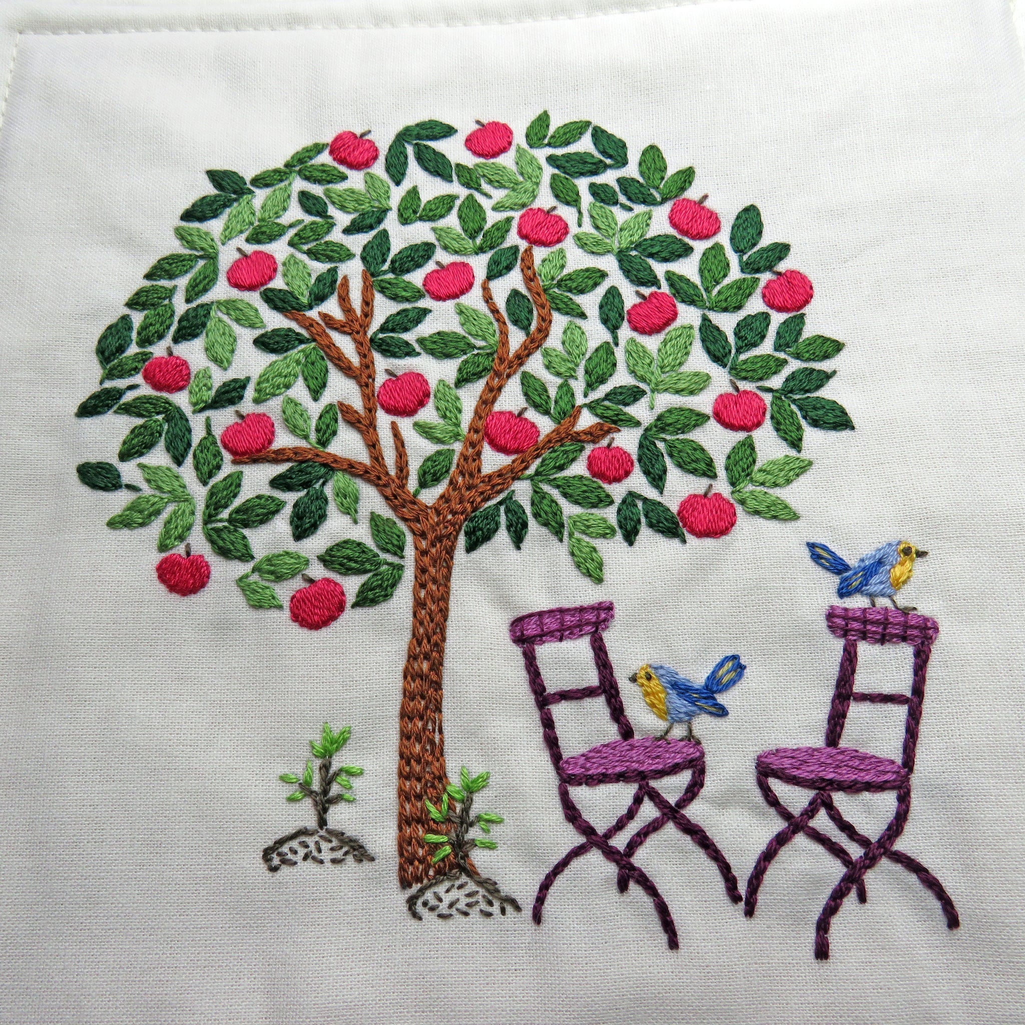 Embroidered design of a tree with birds and chairs on a white background