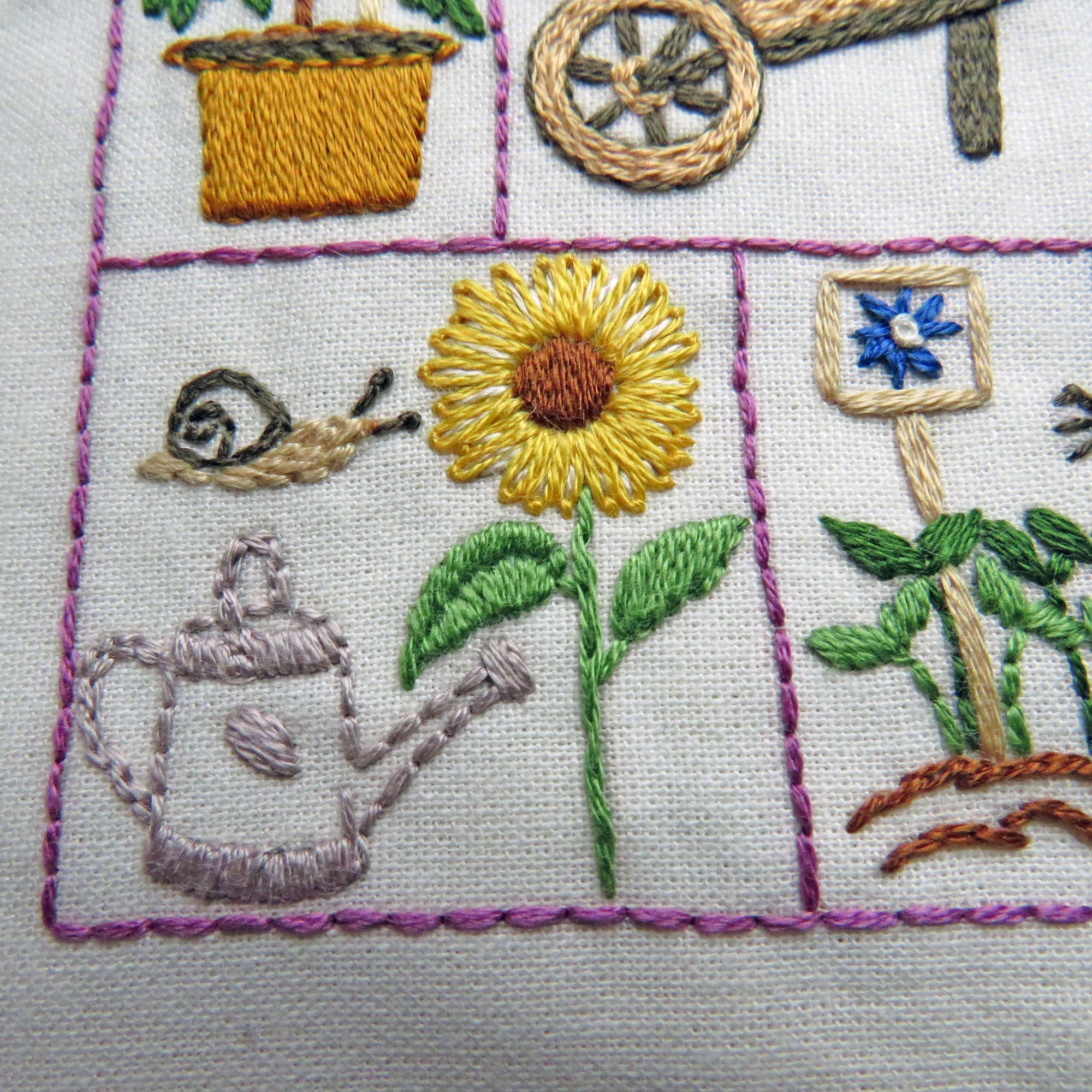 Embroidered panel with garden-themed designs including a sunflower, snail, watering can, and flower.