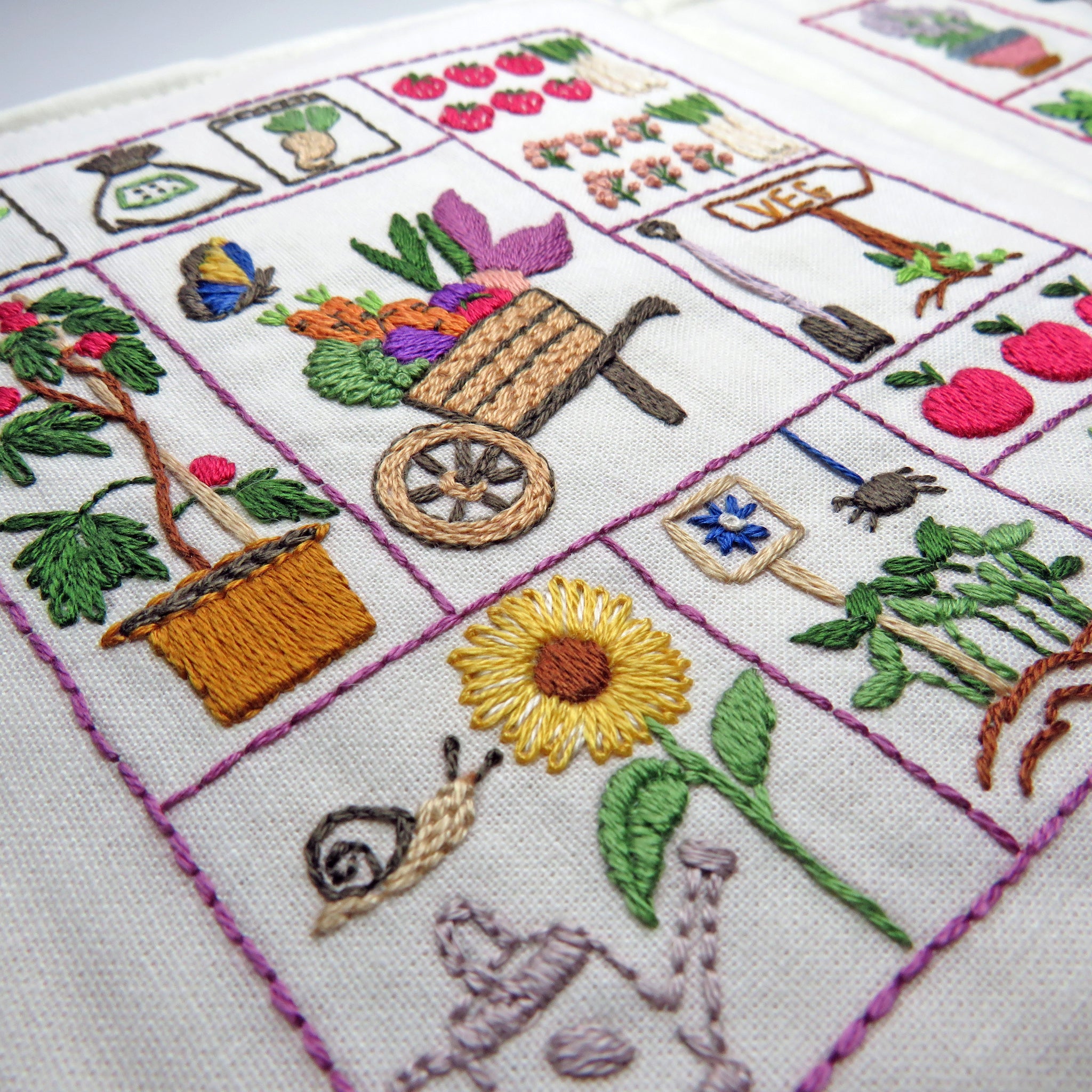 Embroidered garden scene with flowers, a wheelbarrow, and plants on white fabric.