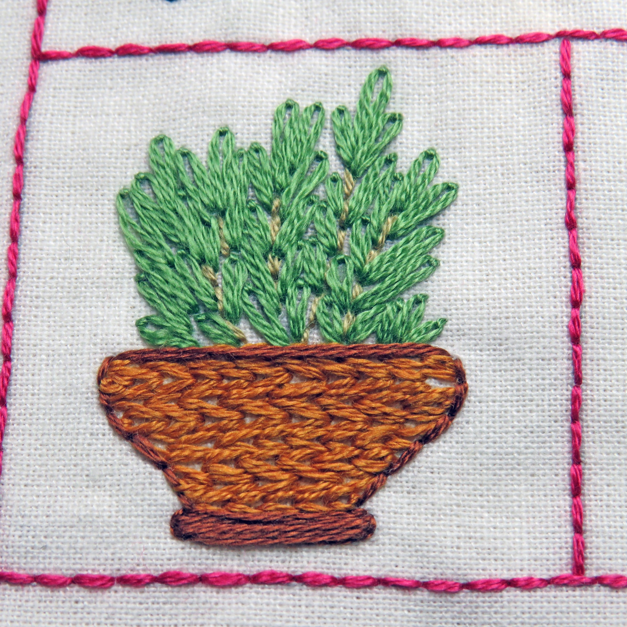 Embroidered potted plant with green leaves and brown pot on white fabric with pink stitching.