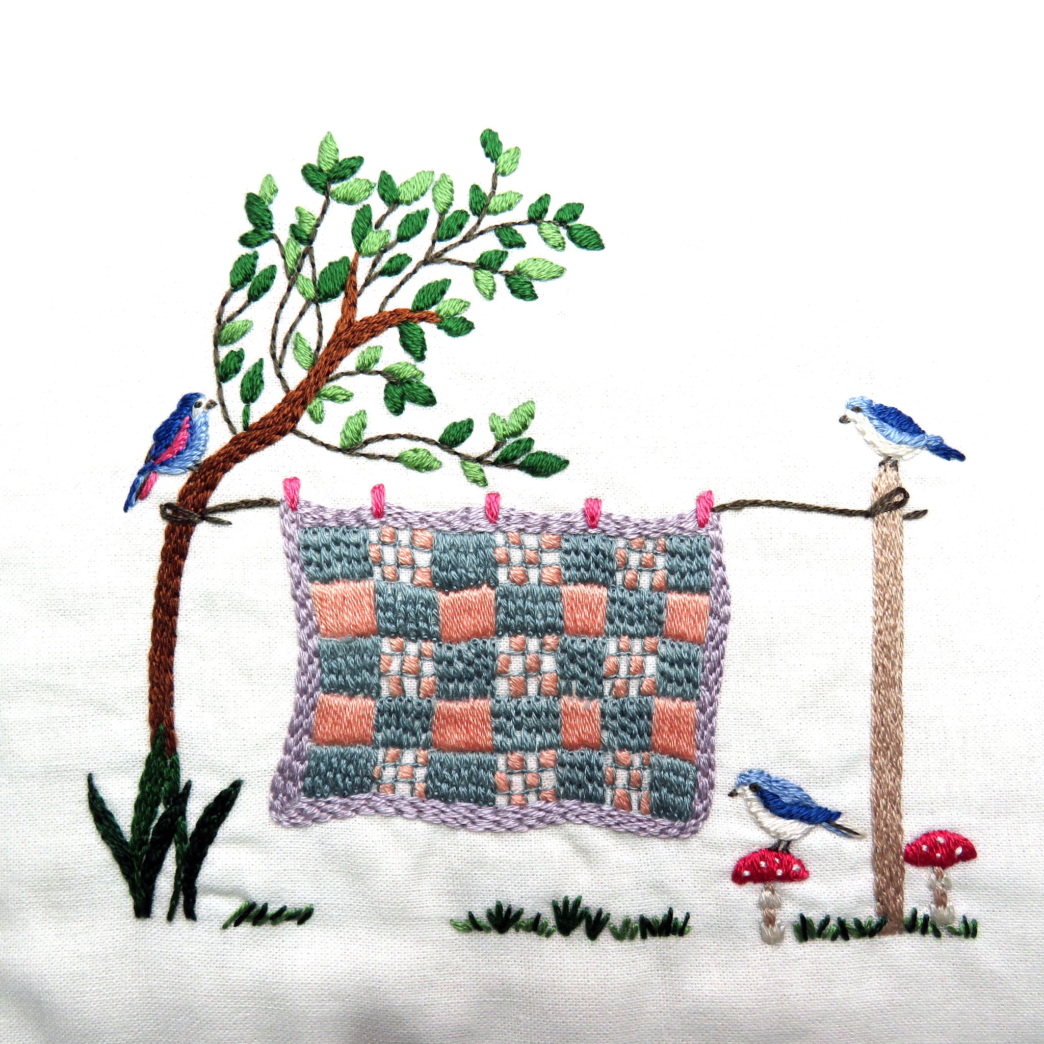 Embroidered scene with a checkered quilt, birds, and a tree on a white background