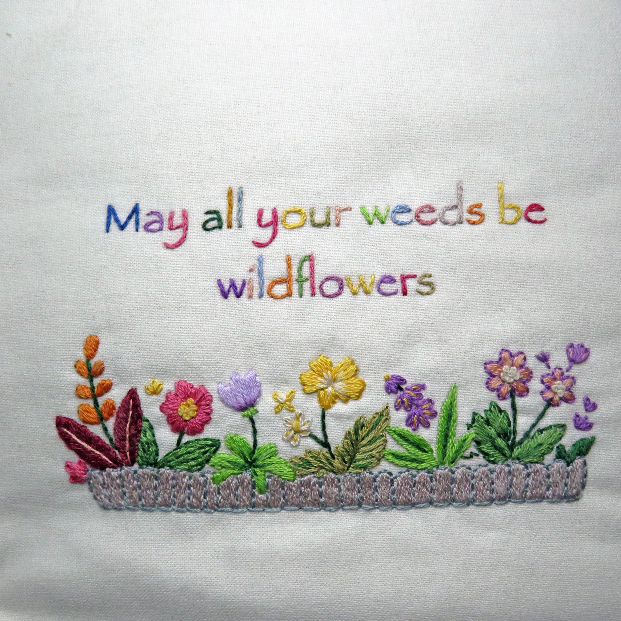 Embroidered text 'May all your weeds be wildflowers' with colorful flowers on a white background