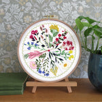 Embroidery hoop with floral design on a wooden stand against a decorative wall.