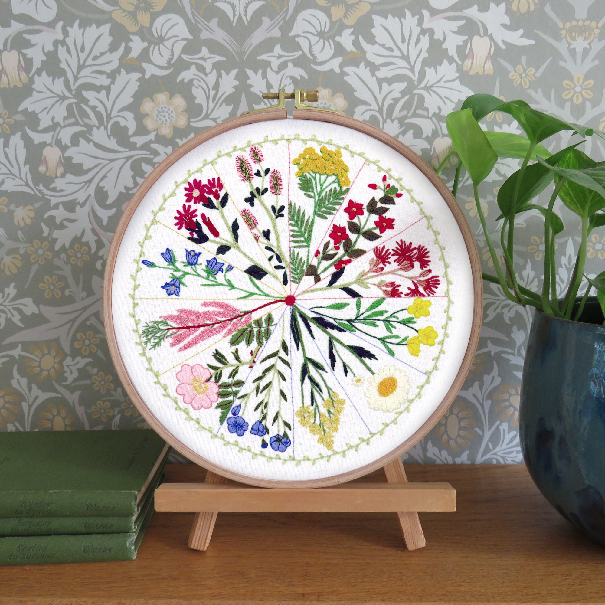 Embroidery hoop with floral design on a wooden stand against a decorative wall.