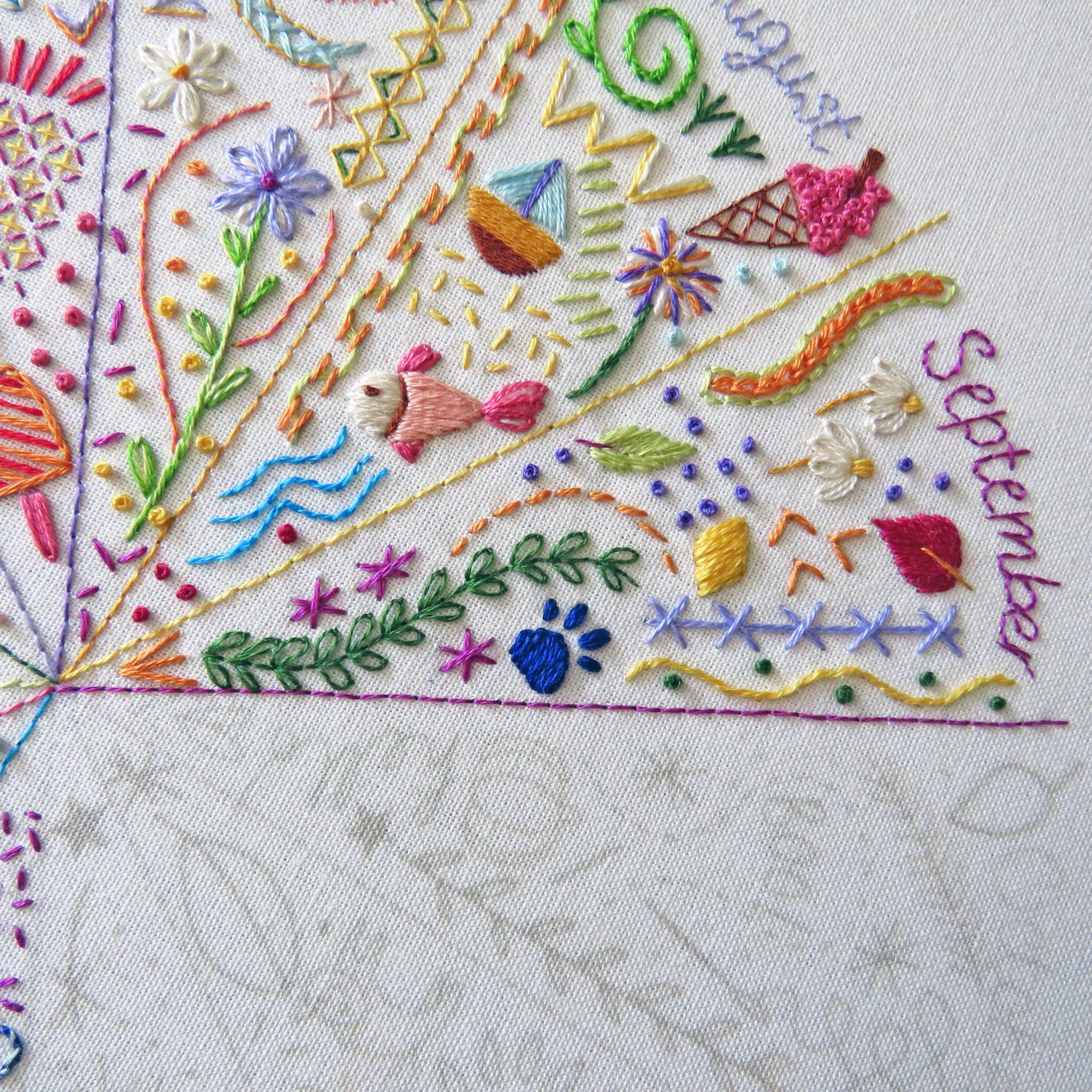 Close-up of a colorful embroidered fabric design featuring seasonal motifs like a fish, flowers, leaves, and handwritten text reading 'August' and 'September' on a white textile background.
