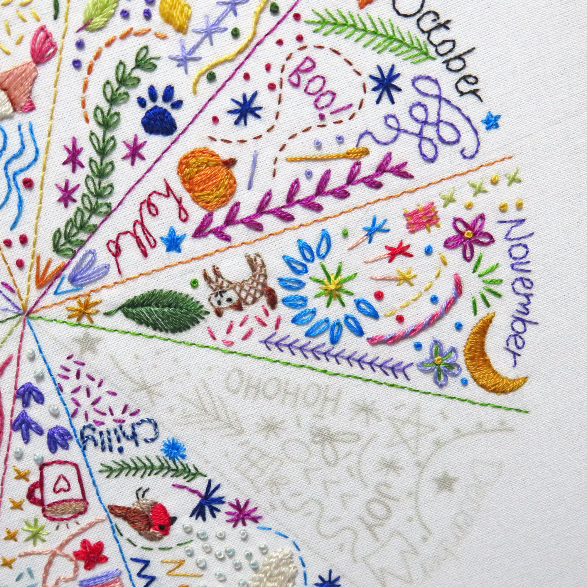 Close-up of a white fabric surface with colorful embroidered seasonal sections and text reading 'October', 'November', and 'December', plus decorative motifs like leaves, stars, flowers, a paw print, a bird, and a small dog.