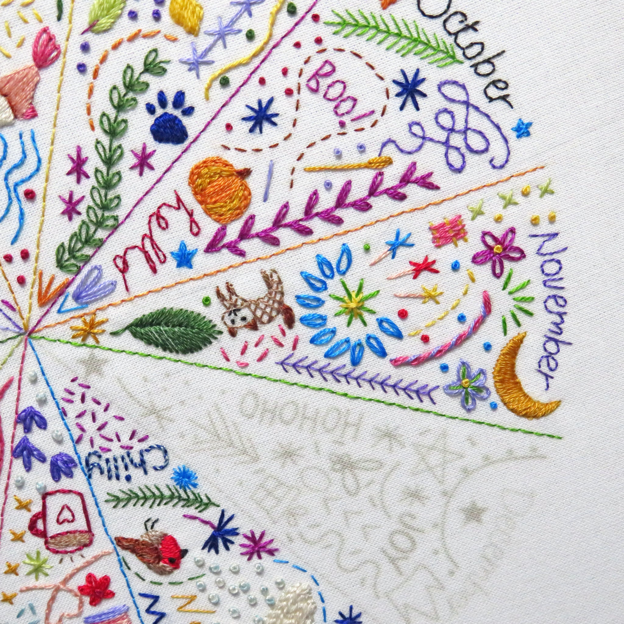 Close-up of a white fabric surface with colorful embroidered seasonal sections and text reading 'October', 'November', and 'December', plus decorative motifs like leaves, stars, flowers, a paw print, a bird, and a small dog.