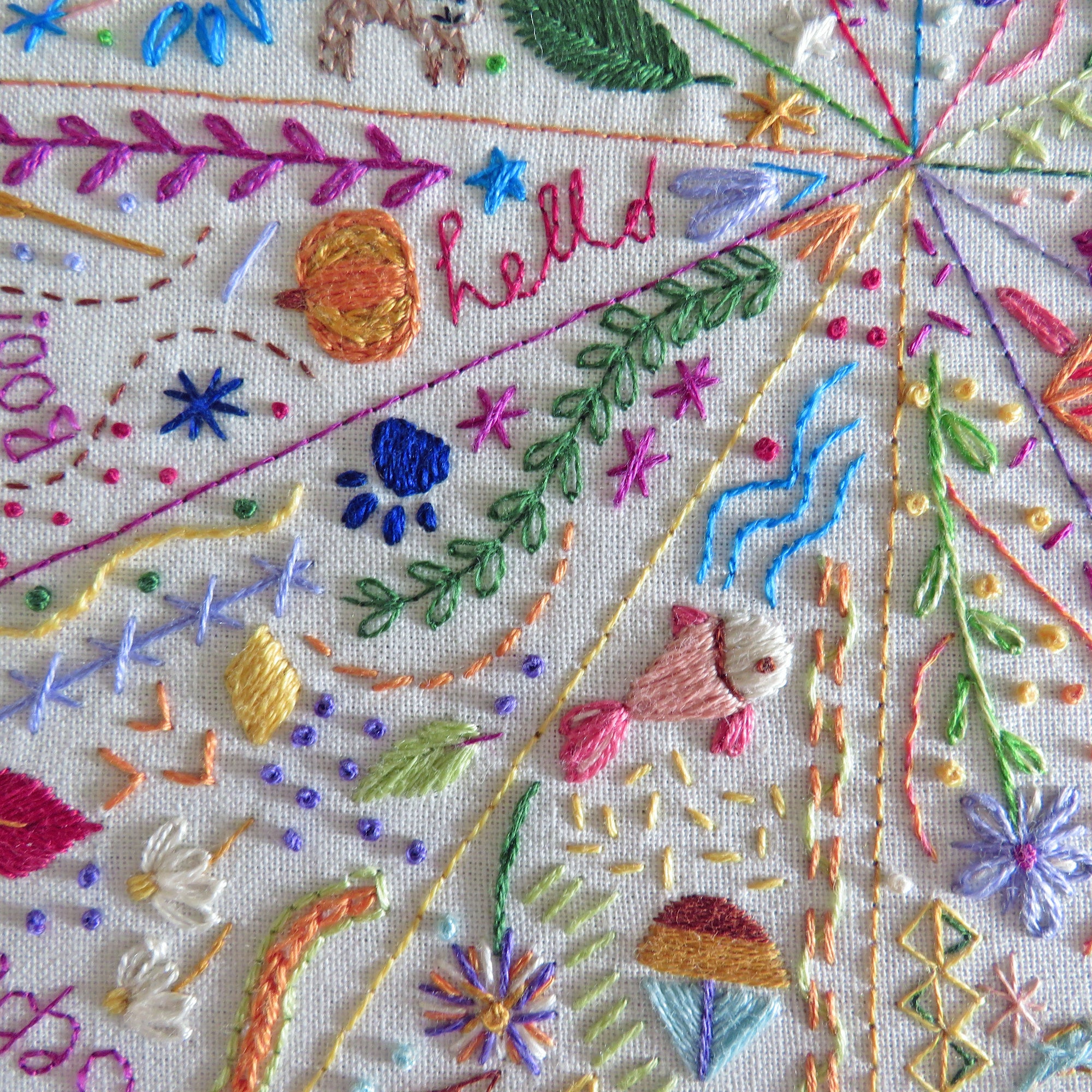 Close-up of a colorful embroidered textile featuring floral motifs, leaves, geometric patterns, and the word "hello" stitched in multicolored thread on a cream fabric background.