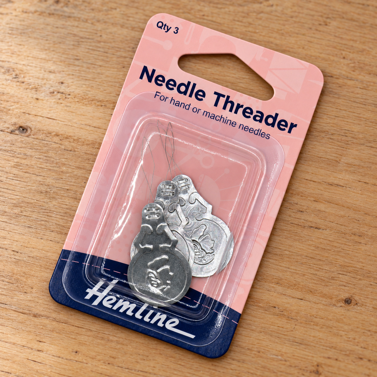 A packaged Hemline needle threader set on a wooden surface. The product consists of two silver metal threaders (one for hand/machine needles) in a clear plastic blister pack attached to a pink and navy blue cardboard backing.