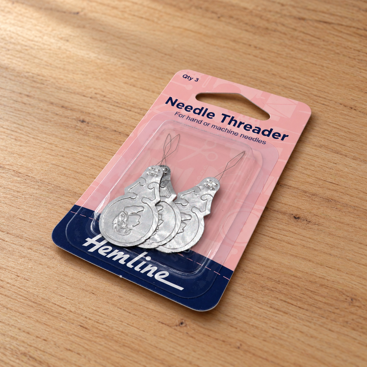 A pair of silver metal needle threaders in blister packaging on a light wood surface. The backing card is pink and navy blue with the brand name 'Hemline' visible.