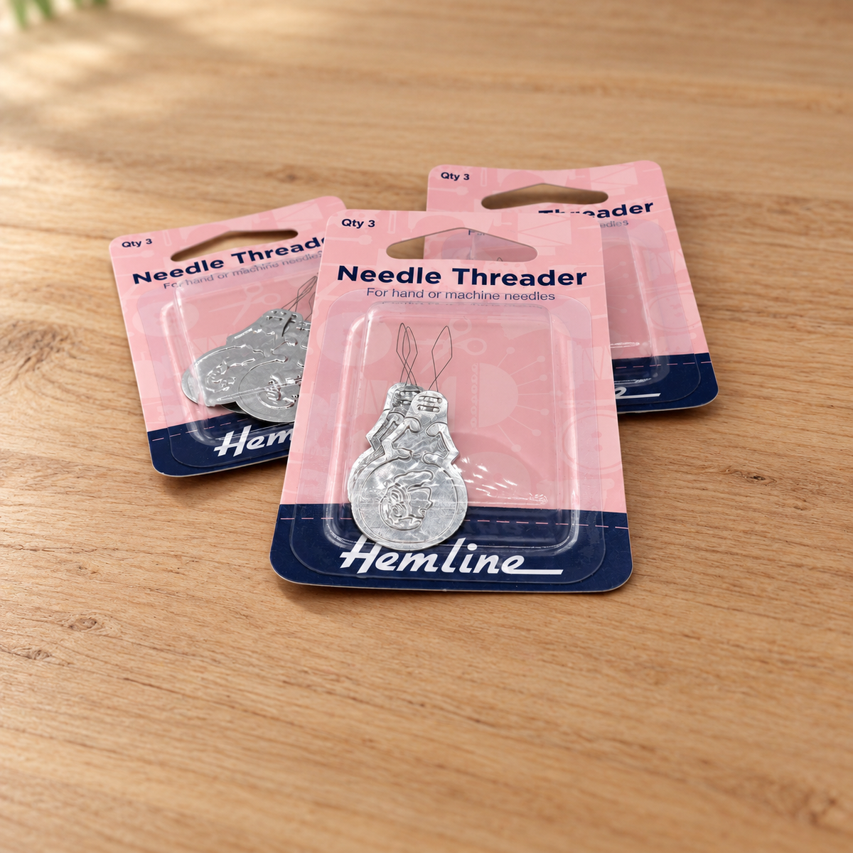 Three packaged Hemline needle threaders arranged on a wooden tabletop. Each item is in a pink and navy blister pack with a silver metal thread holder visible inside.