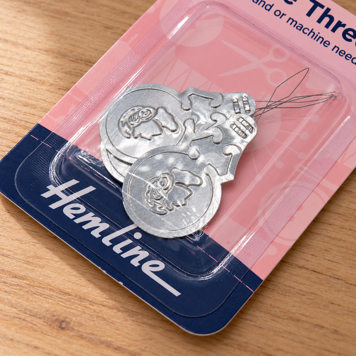 Silver metal thread spools in a pink and navy blue Hemline branded blister pack, placed on a wooden surface.