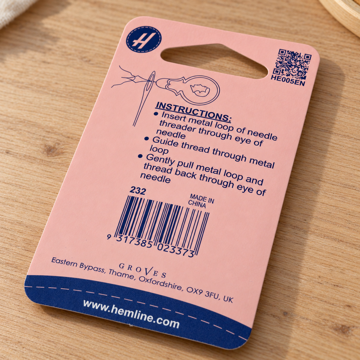 A pink and blue retail card for a hemstitch needle from the brand Groves, placed on a light wooden surface. The card features product instructions, a barcode, a QR code, and branding.