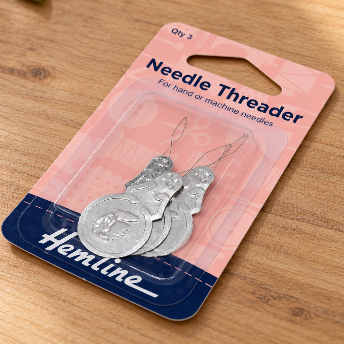 Three silver metal Hemline needle threaders in a pink and blue blister pack on a wooden surface.