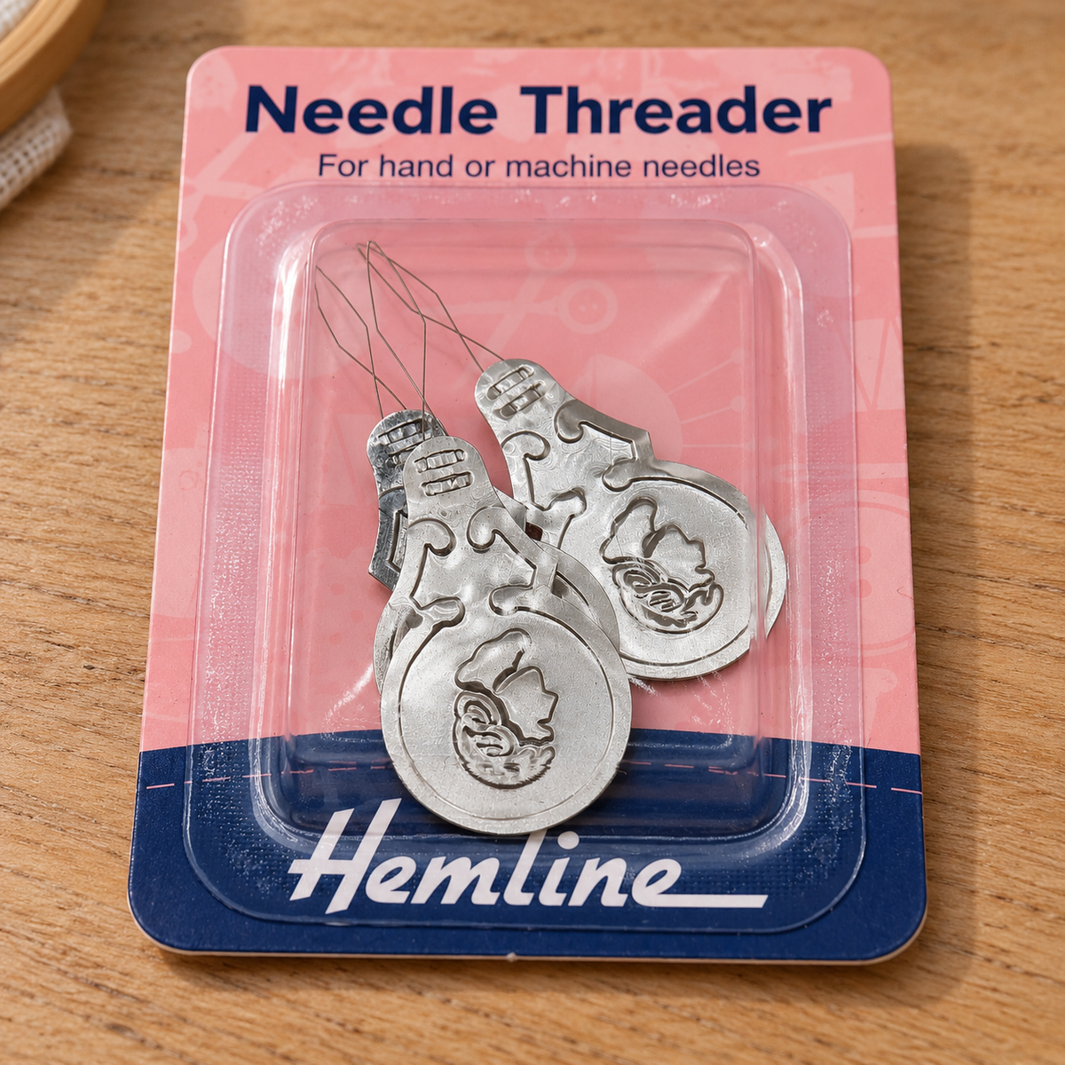 A package of Hemline needle threaders for hand or machine needles, shown in its pink and blue blister packaging on a wooden surface.