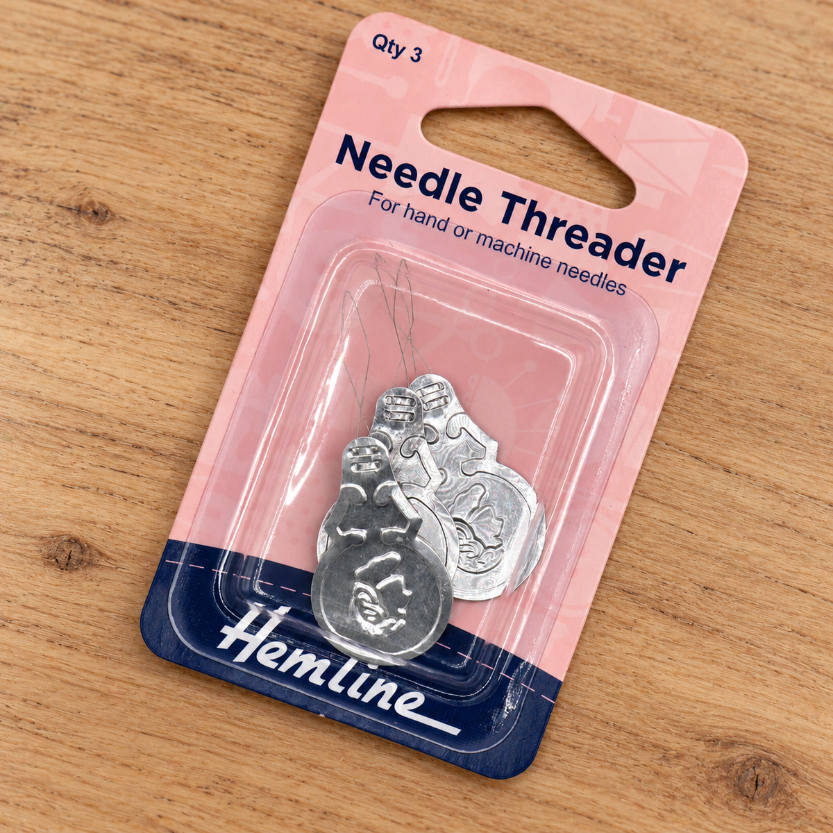 A Hemline Needle Threader in silver metal, packaged in a pink and navy blue blister card on a wooden surface.