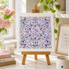 Decorative scene with a colorful floral mandala on an easel, books, flowers, and a mug.