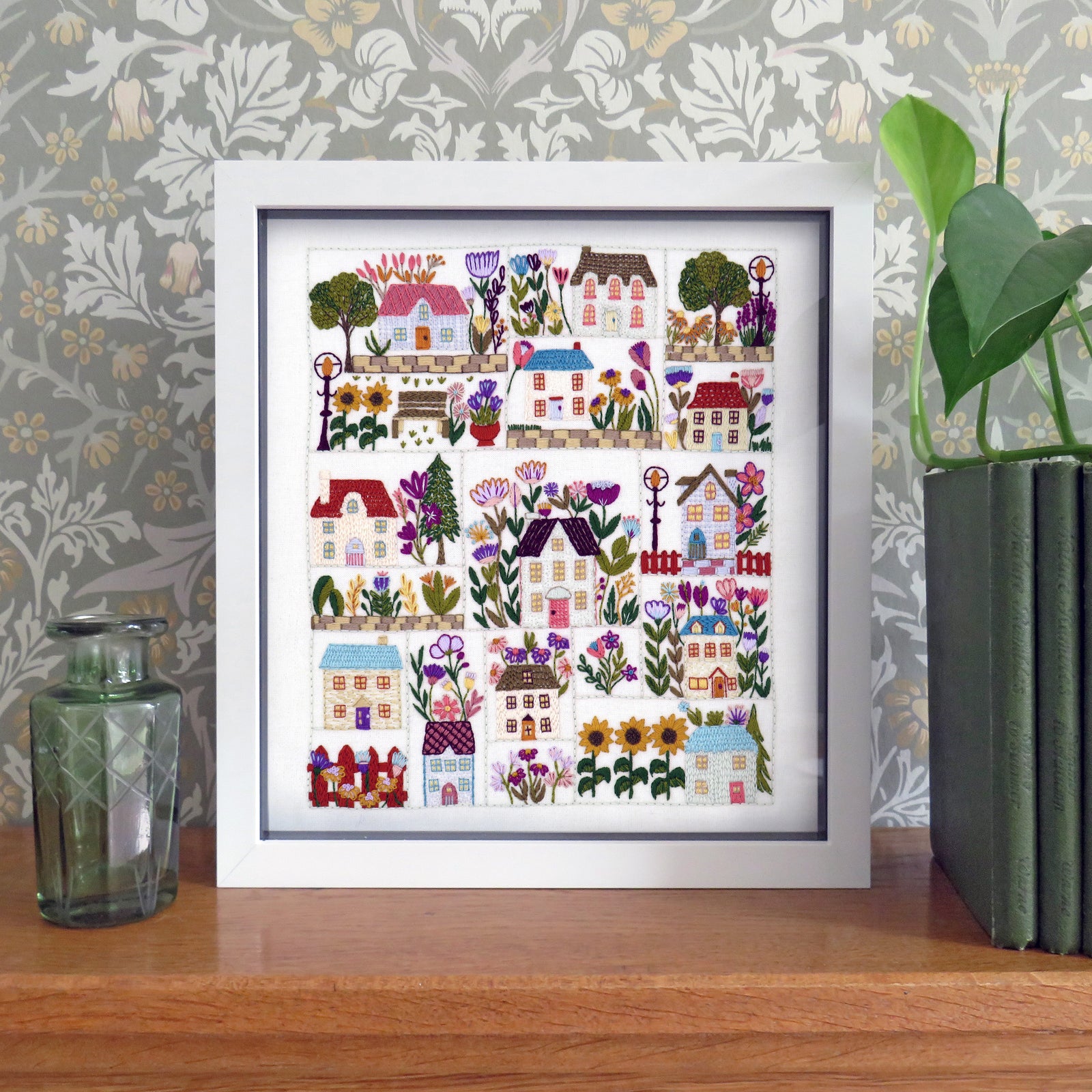 Framed artwork with colorful houses and plants on a wooden surface with decorative wallpaper.
