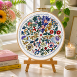 William morris Embroidery hoop with floral design on a desk with books, flowers, and a cup.