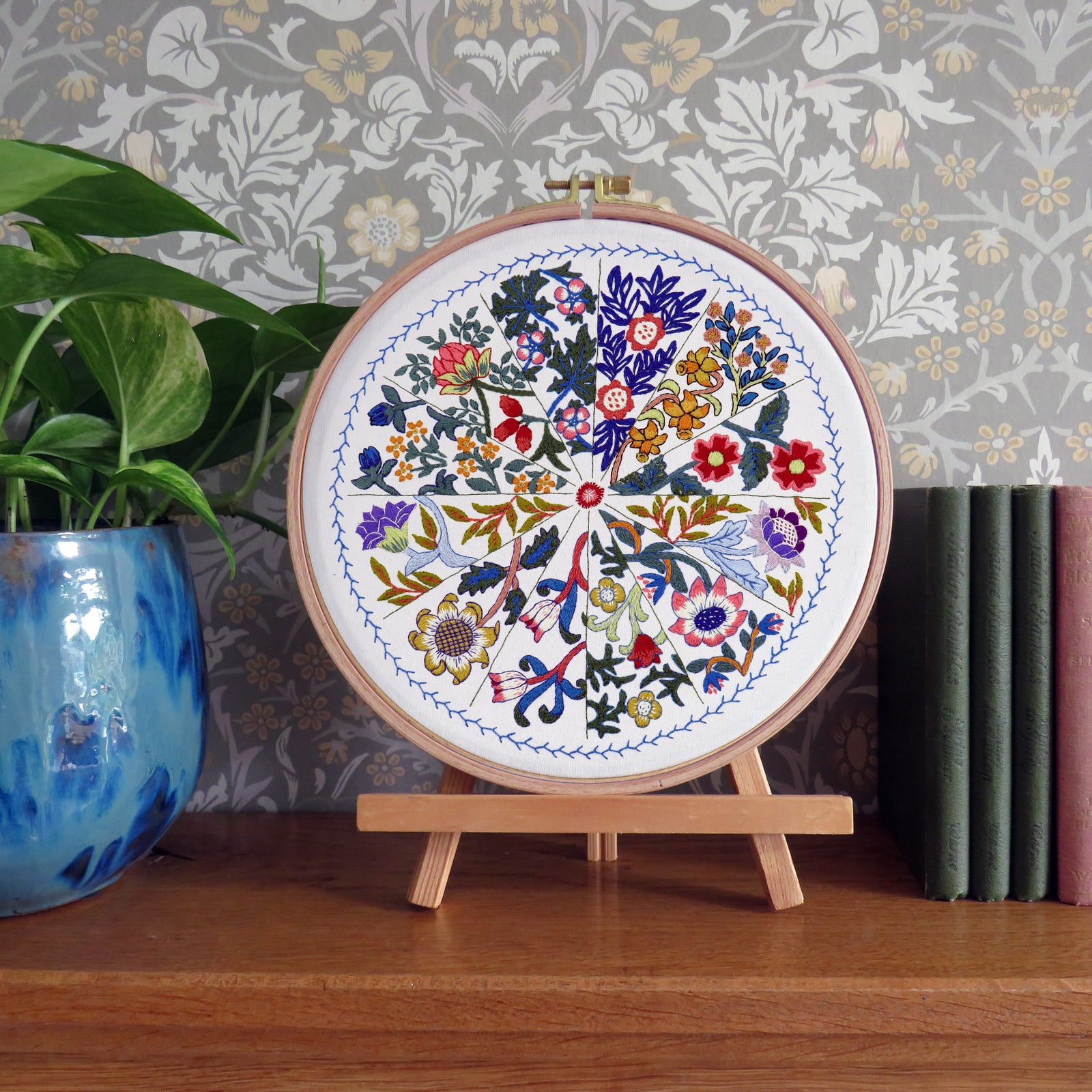 Embroidery hoop with floral pattern on a wooden stand against a decorative wall.