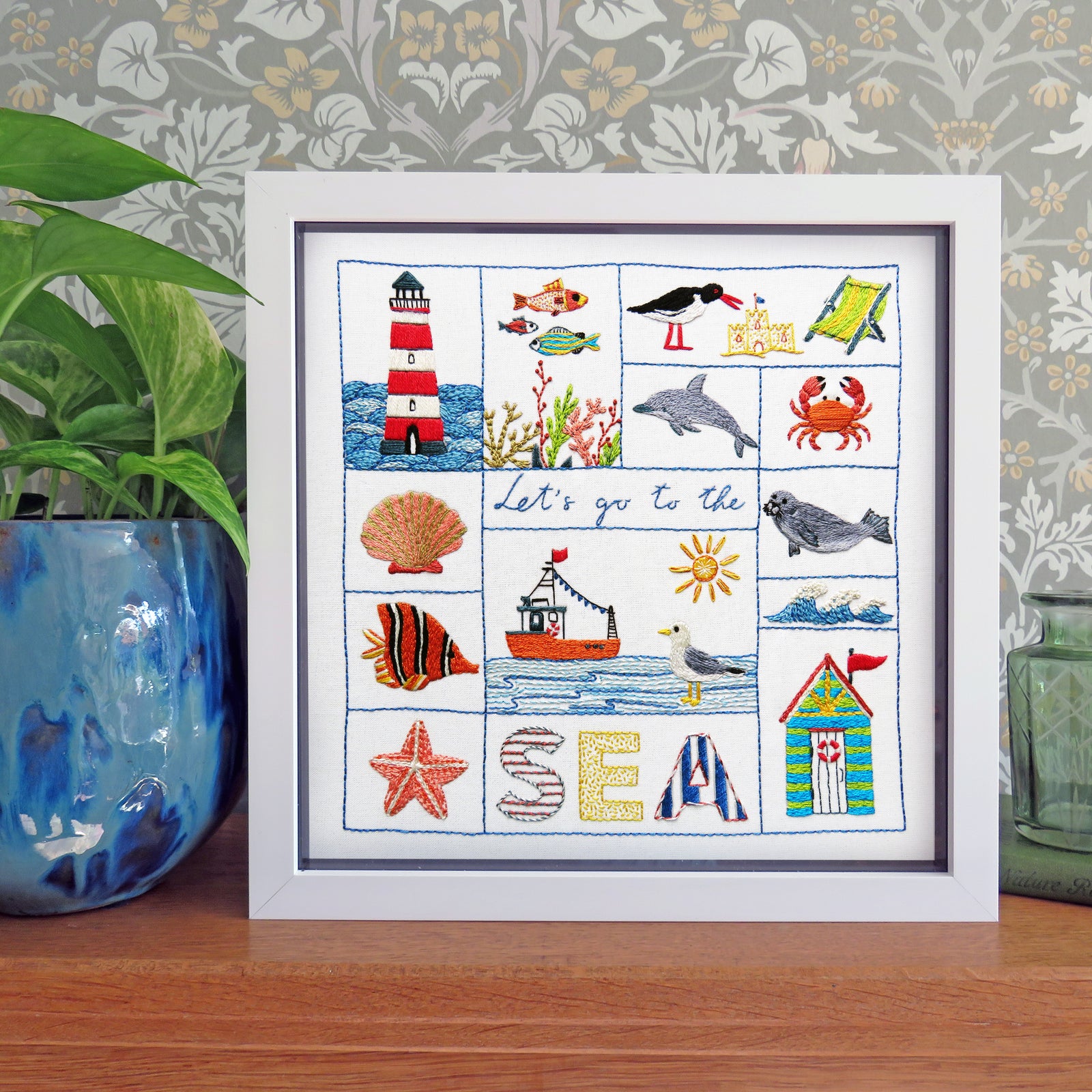 A framed cross-stitch artwork featuring nautical and beach-themed motifs such as a lighthouse, fish, seashells, a boat, a beach house, and the word 'SEA' in decorative lettering. The white-framed piece is displayed on a wooden surface against a patterned wallpaper background, with a blue potted plant on the left and a green container on the right.